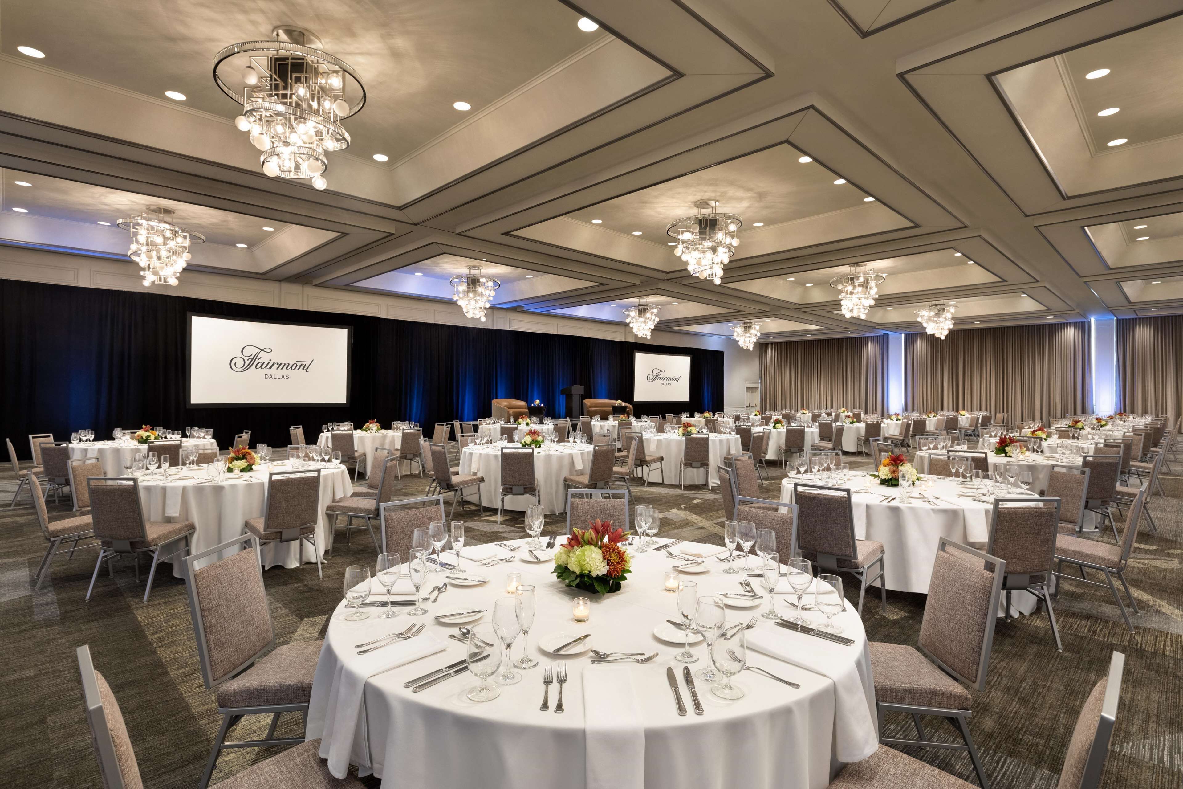 A banquet hall with round tables set for dining, chandeliers, and a projection screen displaying the hotel name.