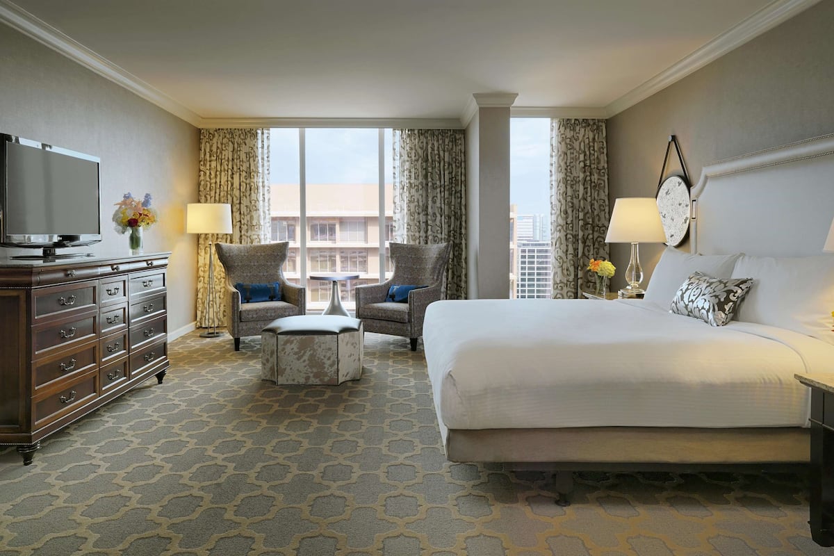 A hotel room with a large bed, a TV, a dresser, two armchairs, a coffee table, and a view of the city.
