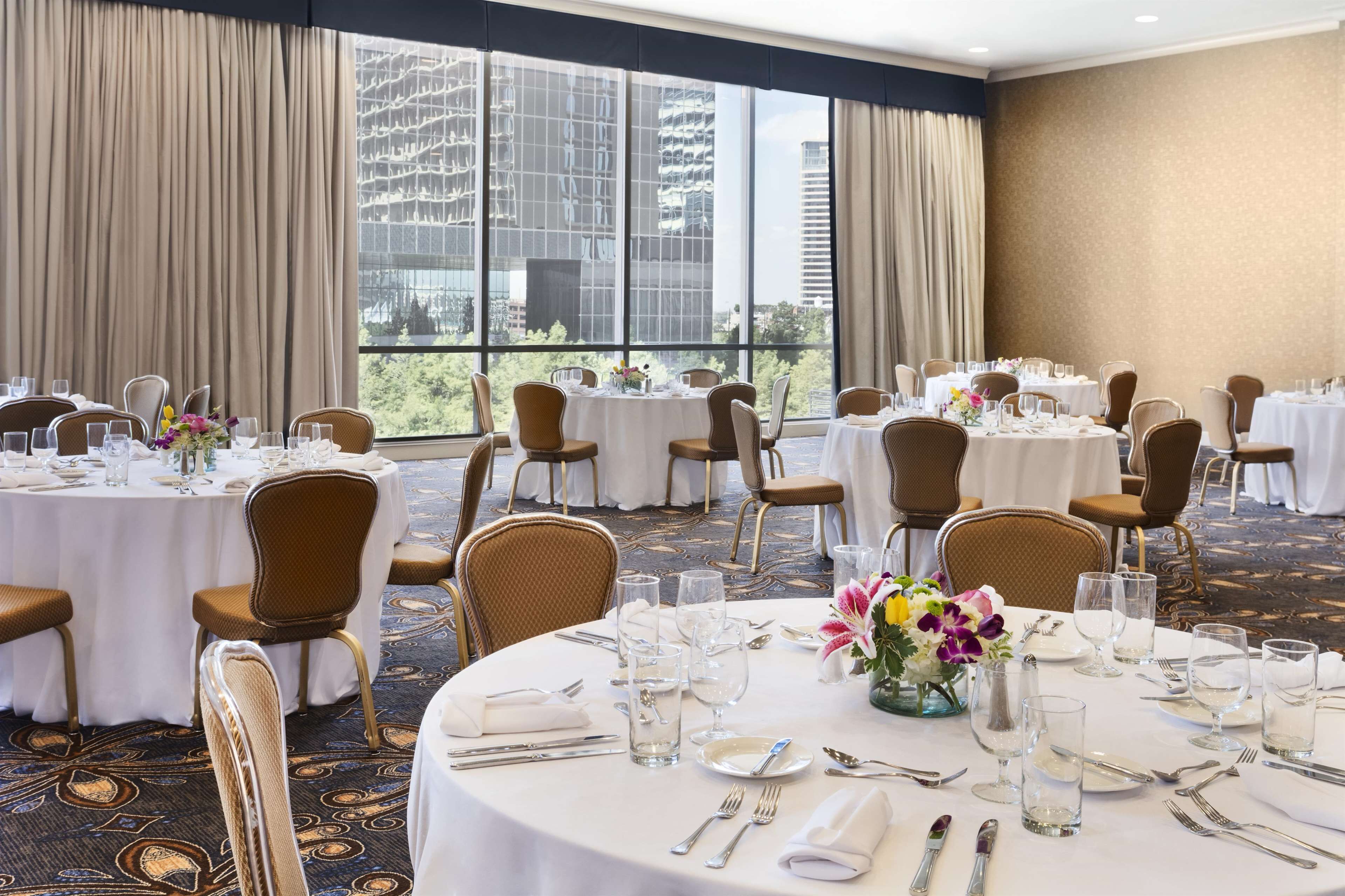Une salle à manger d’hôtel avec des tables rondes dressées pour les clients, de grandes fenêtres offrant une vue sur la ville et un sol recouvert de moquette.