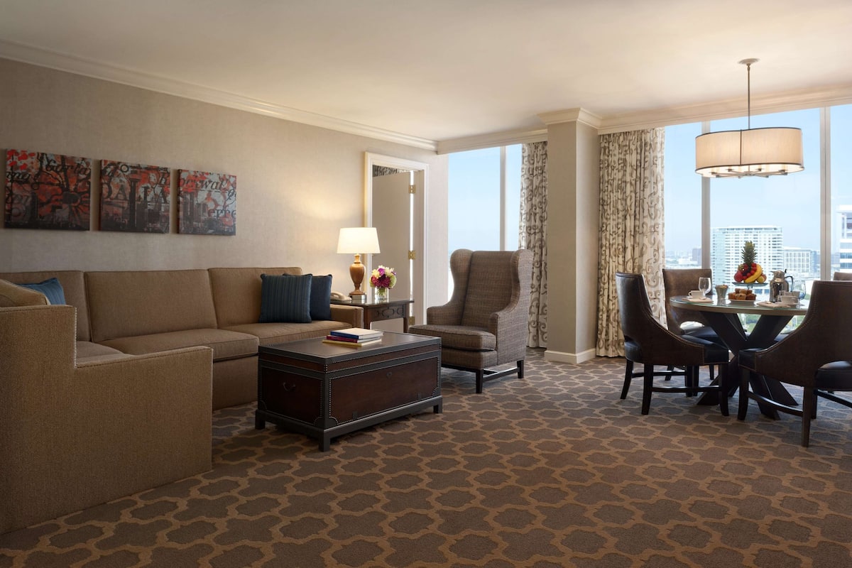 A hotel room with a sofa, armchair, dining table with chairs, and a large window with a city view.