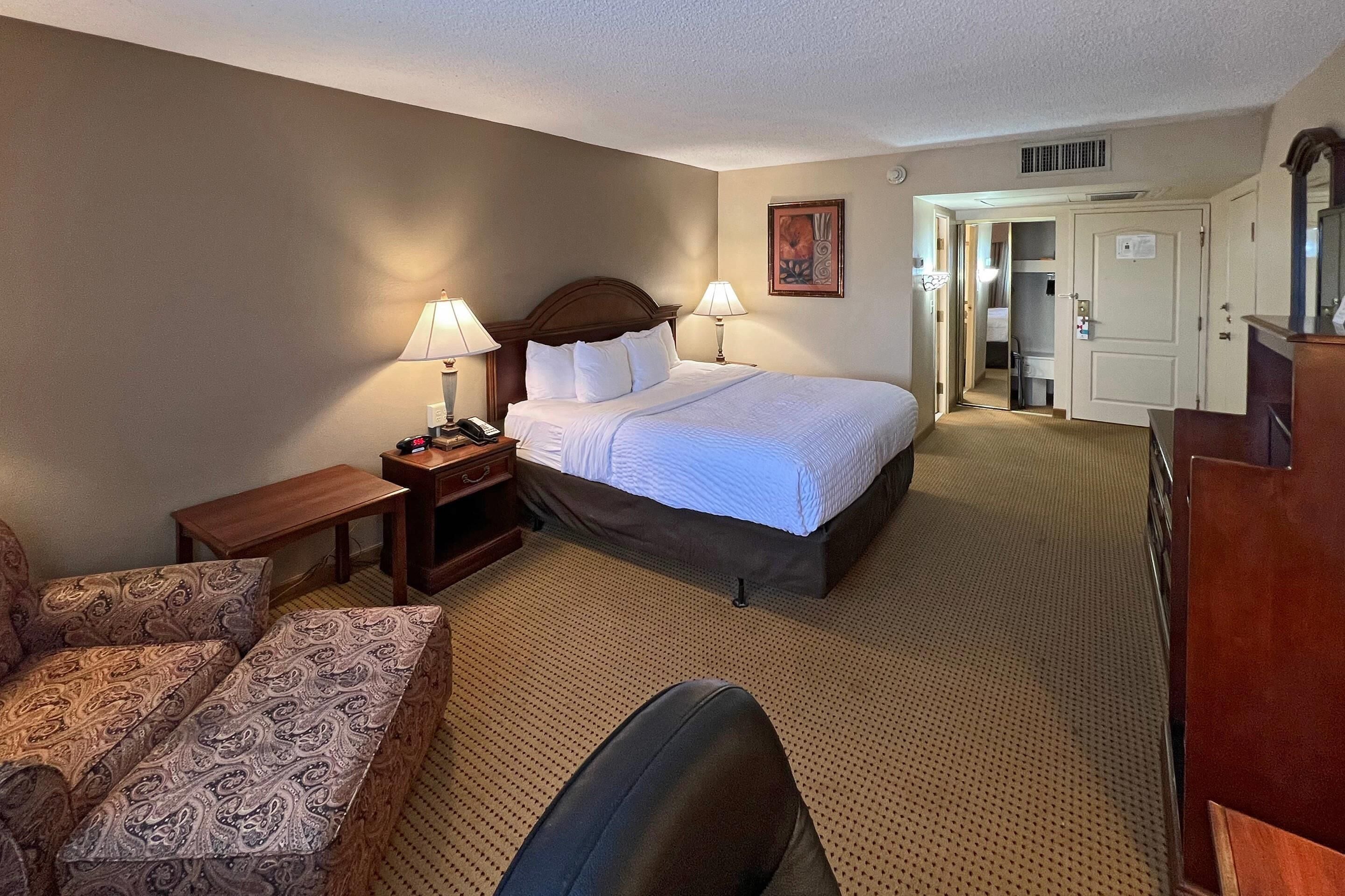 A hotel room with a bed, two bedside lamps, a wooden nightstand, a patterned sofa, a chair, and a desk.