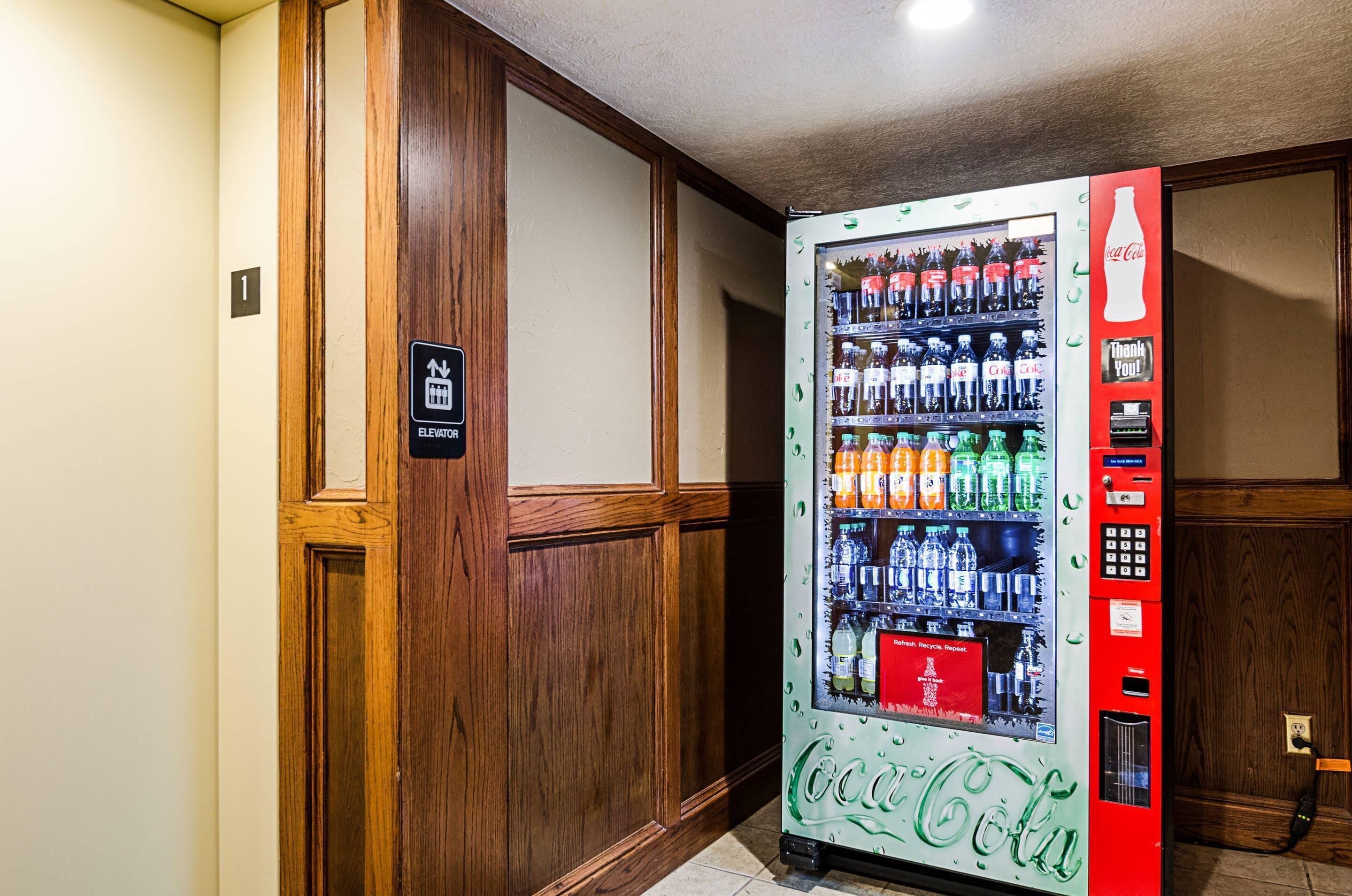 A Coca-Cola vending machine in a hotel lobby with a wooden panel wall and an elevator sign.