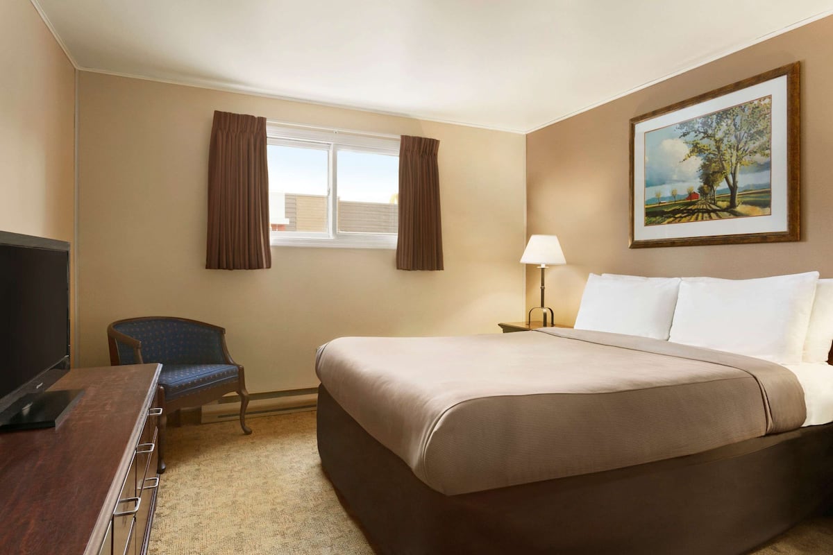 A hotel room with a bed, a desk with a TV, a chair, a window with curtains, and a painting on the wall.