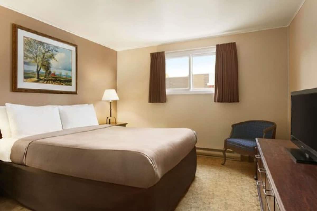 A hotel room with a bed, a chair, a desk with a television, a window with curtains, and a painting on the wall.