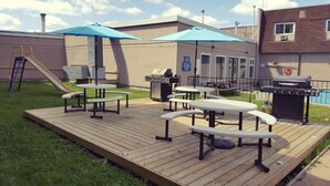 Un patio trasero con una terraza de madera, mesas de picnic, una parrilla y un tobogán.
