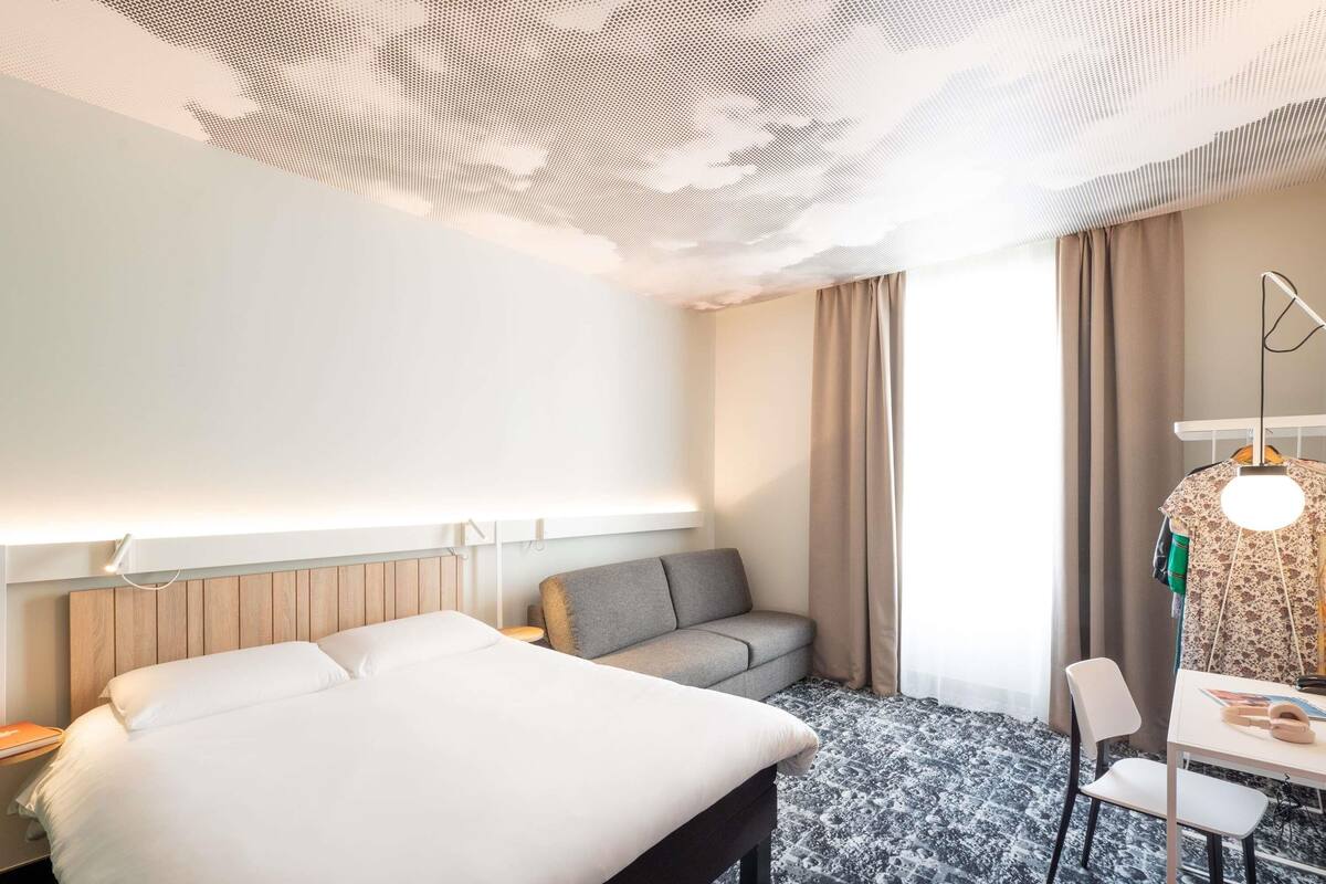 Ett hotellrum med en säng, en soffa, ett skrivbord med en stol, en hängande lampa och ett fönster med gardiner.