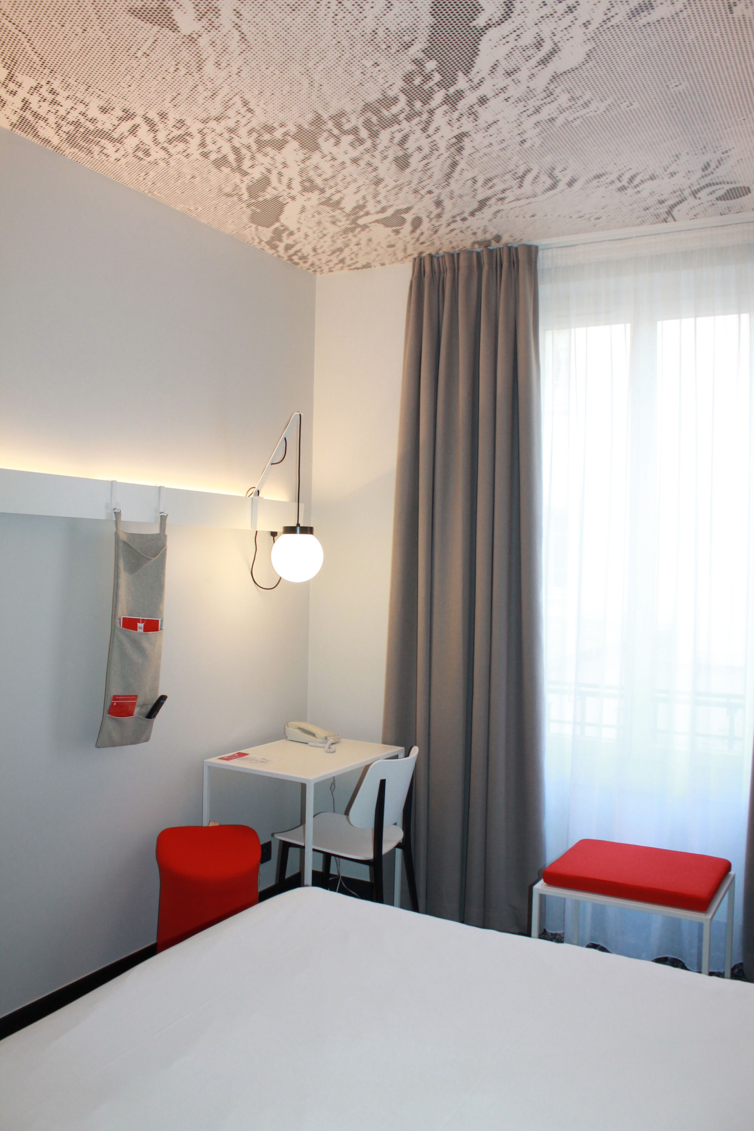 Une chambre d’hôtel avec un lit, un bureau, une chaise, un téléphone, une poubelle rouge et une fenêtre avec des rideaux.