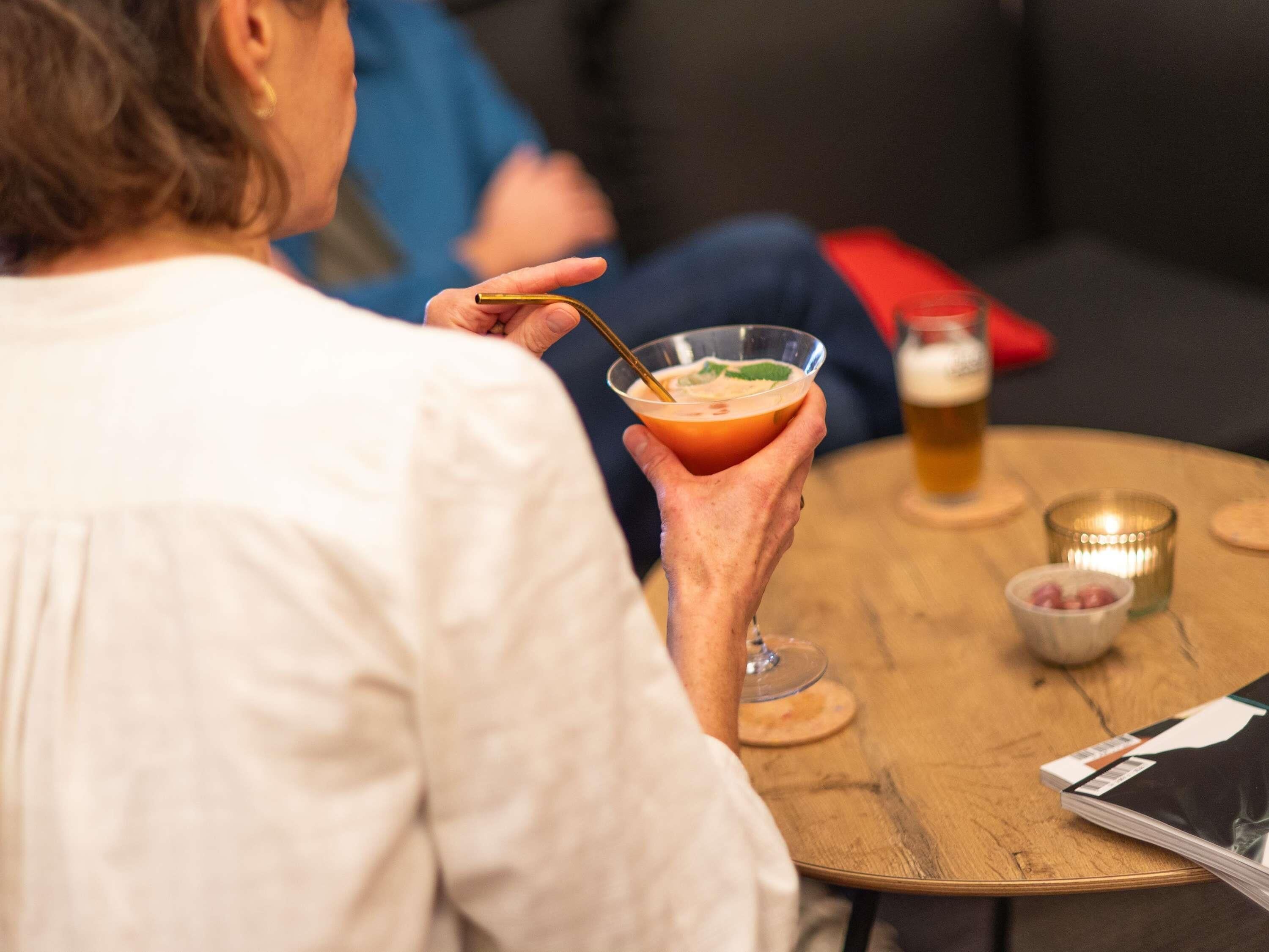 A person holding a cocktail with a straw, seated at a wooden table with a glass of beer and a candle in the background.