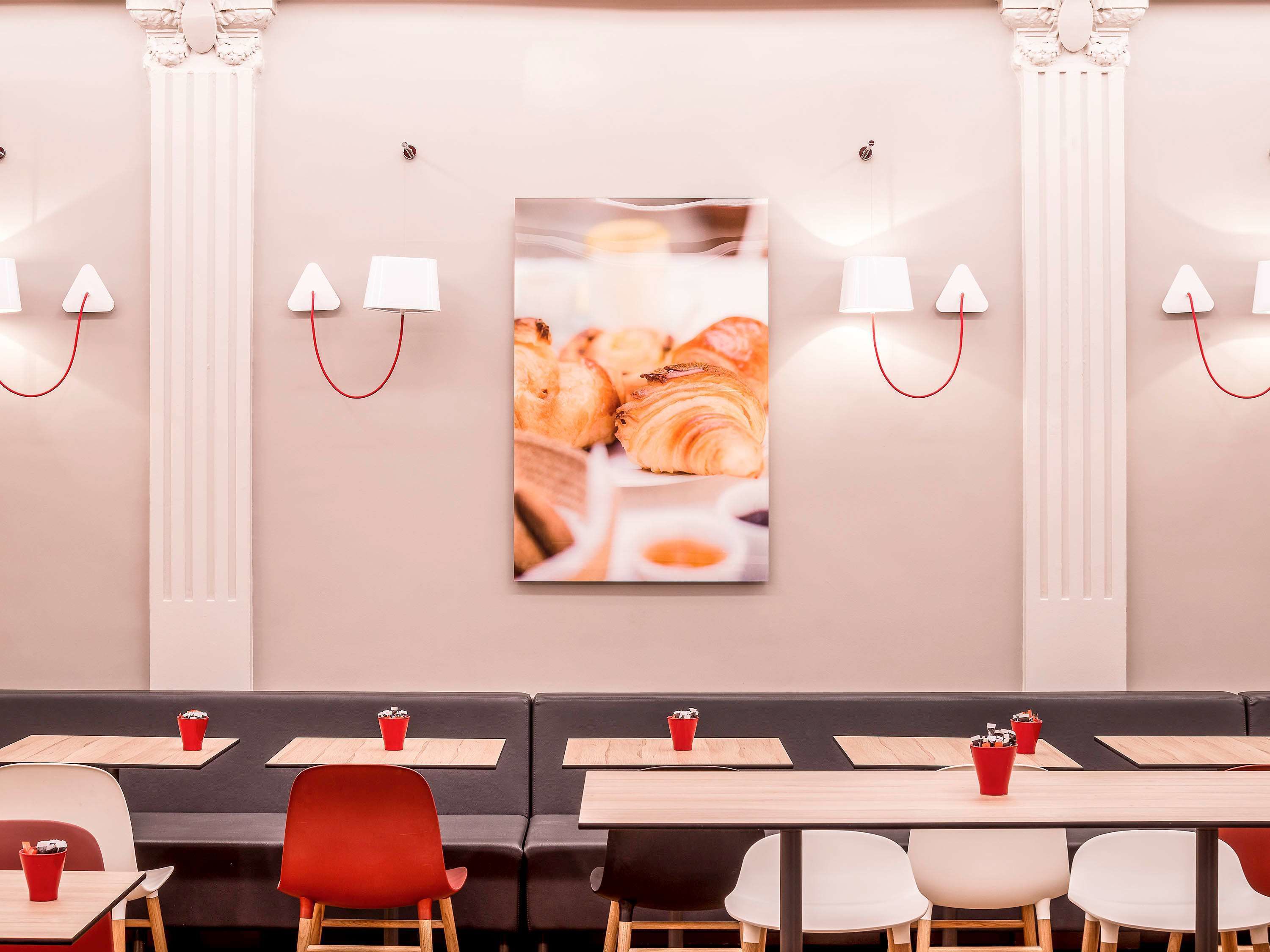 A modern restaurant interior with a central wall-mounted picture of pastries, wooden tables with red cups, and black and white chairs.
