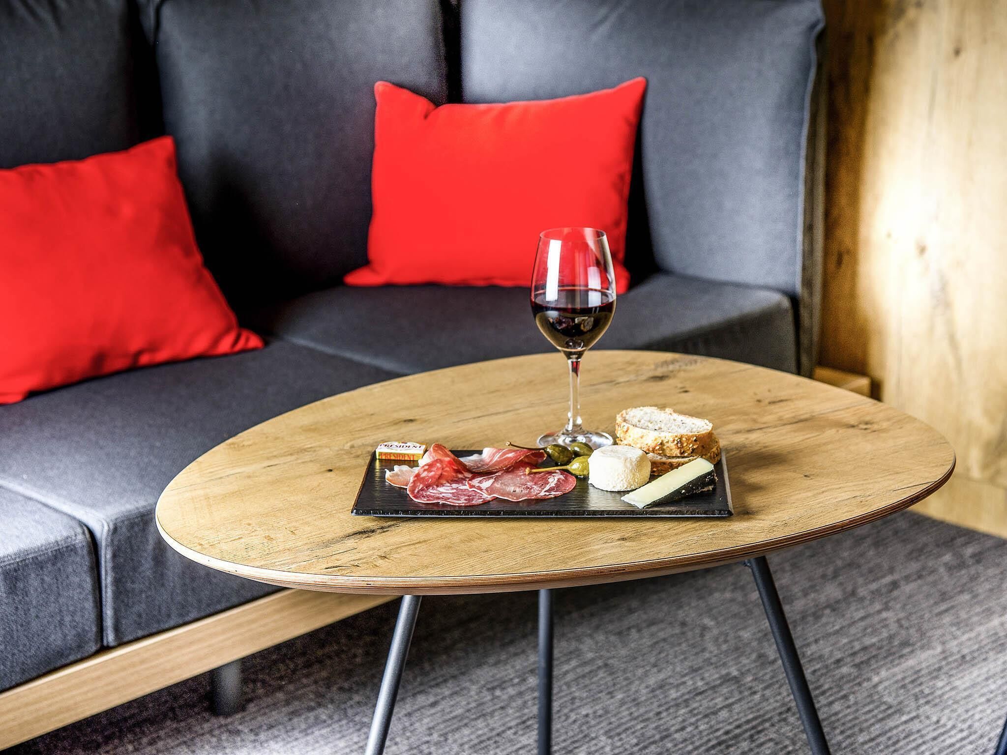A round wooden table with a black slate board holding various cheeses, cured meats, and bread, accompanied by a glass of red wine, set on a grey sofa with red cushions.