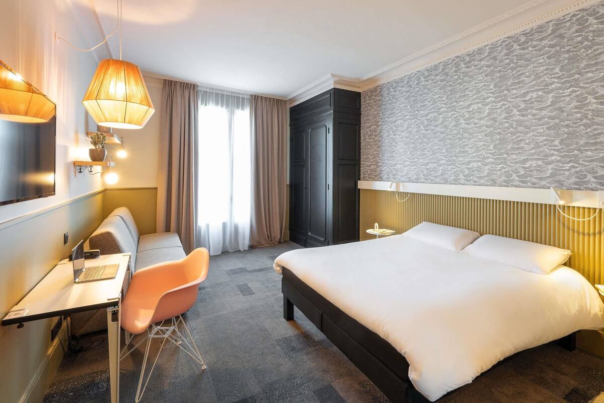 Ett hotellrum med en säng, ett skrivbord, en stol och ett stort fönster med gardiner.
