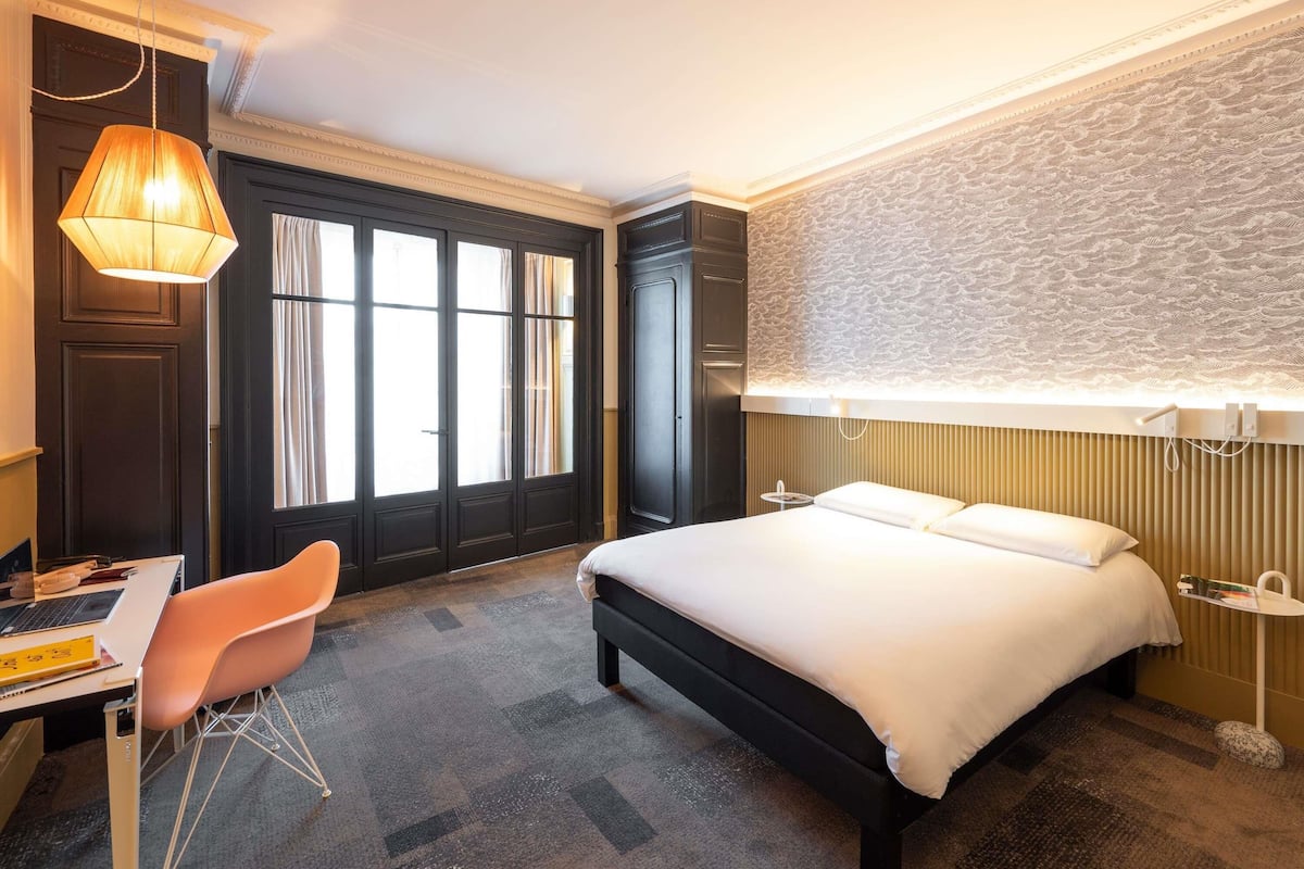 Ett hotellrum med en säng, ett skrivbord, en stol och ett stort fönster med gardiner.