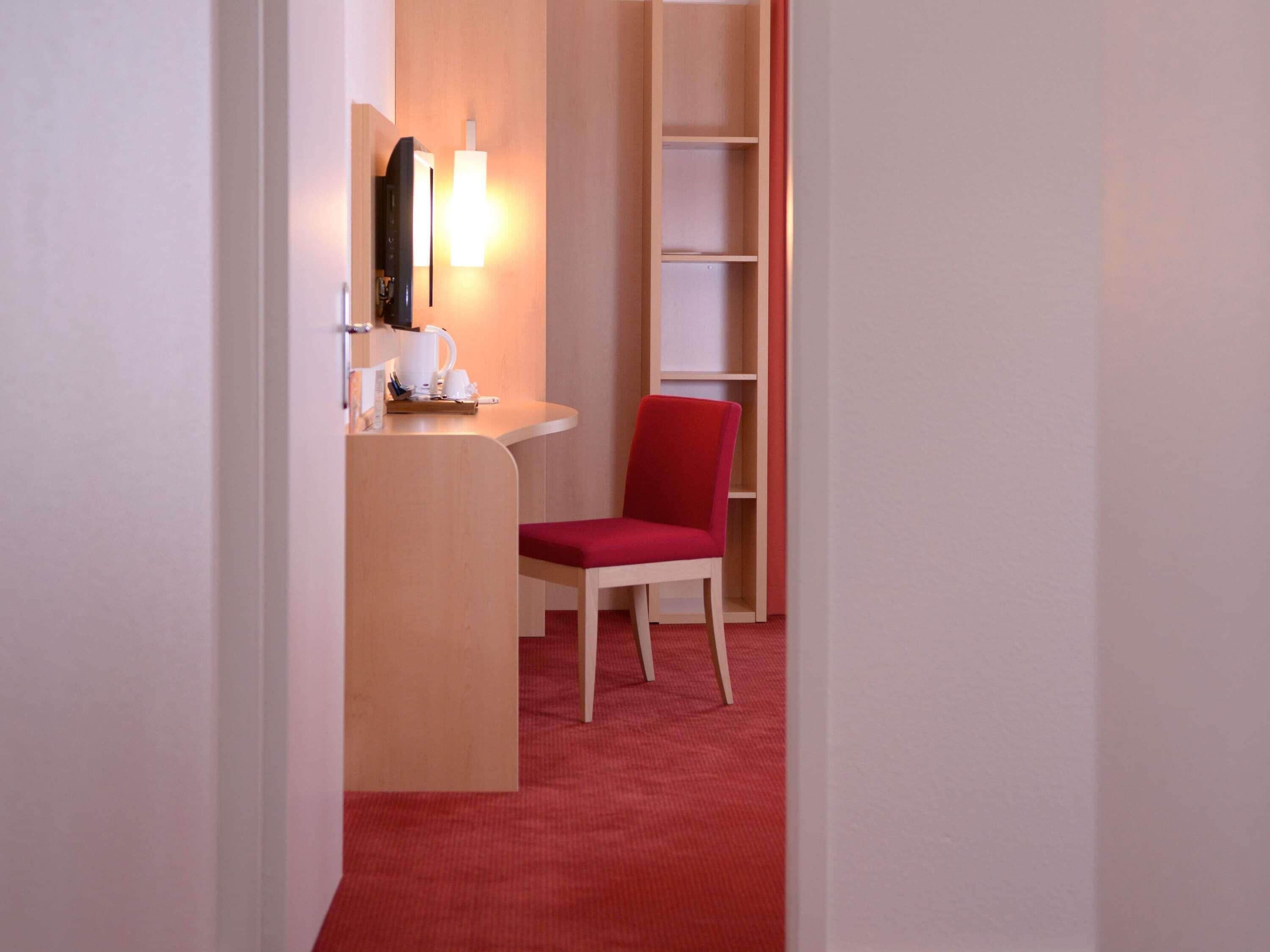 Een hotelkamer met een rood tapijt, een houten bureau met kop en schotel, een rode stoel en een boekenkast.