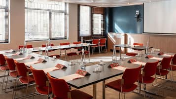 Ein Konferenzraum mit mehreren Tischen und roten Stühlen, großen Fenstern mit Jalousien und einer Projektionsleinwand an der Wand.