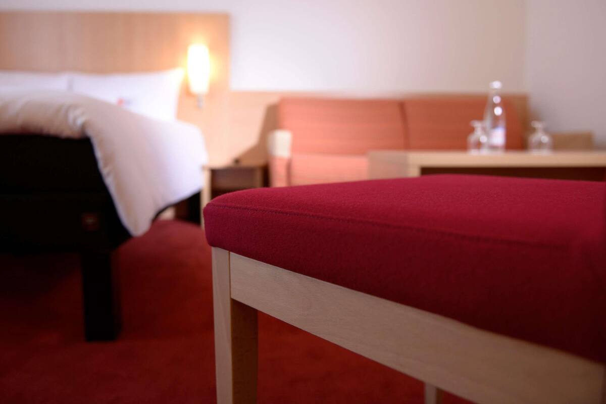 Ett hotellrum med en säng, en röd klädd bänk, ett träbord och en vägglampa.