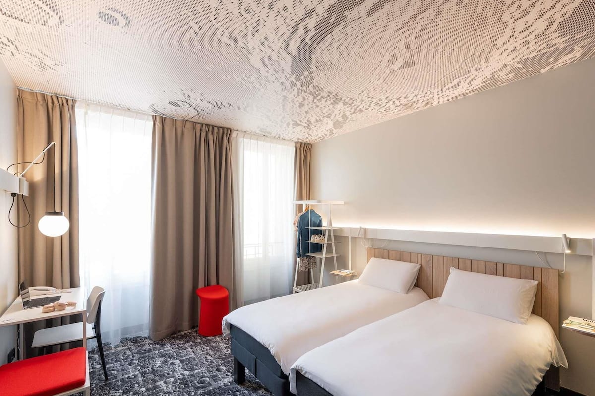 A hotel room with two beds, a desk, a chair, a red stool, and a wardrobe.