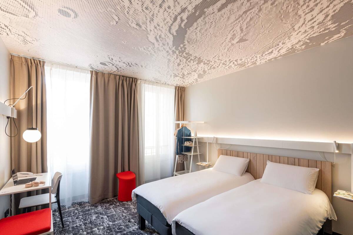Ett hotellrum med två sängar, ett skrivbord, en stol, en röd pall, en garderob och ett mönstrat tak.
