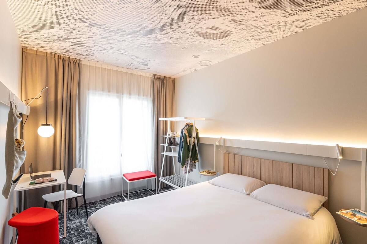 Ett hotellrum med en stor säng, ett skrivbord med en bärbar dator, en röd stol och en klädställning.
