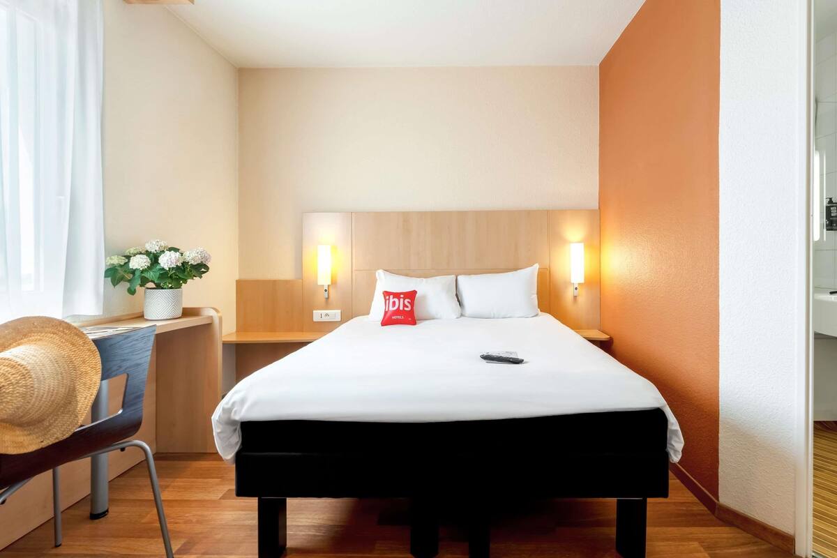 A hotel room with a bed, bedside tables, a desk with a chair, a potted plant, and a wall-mounted light fixture.