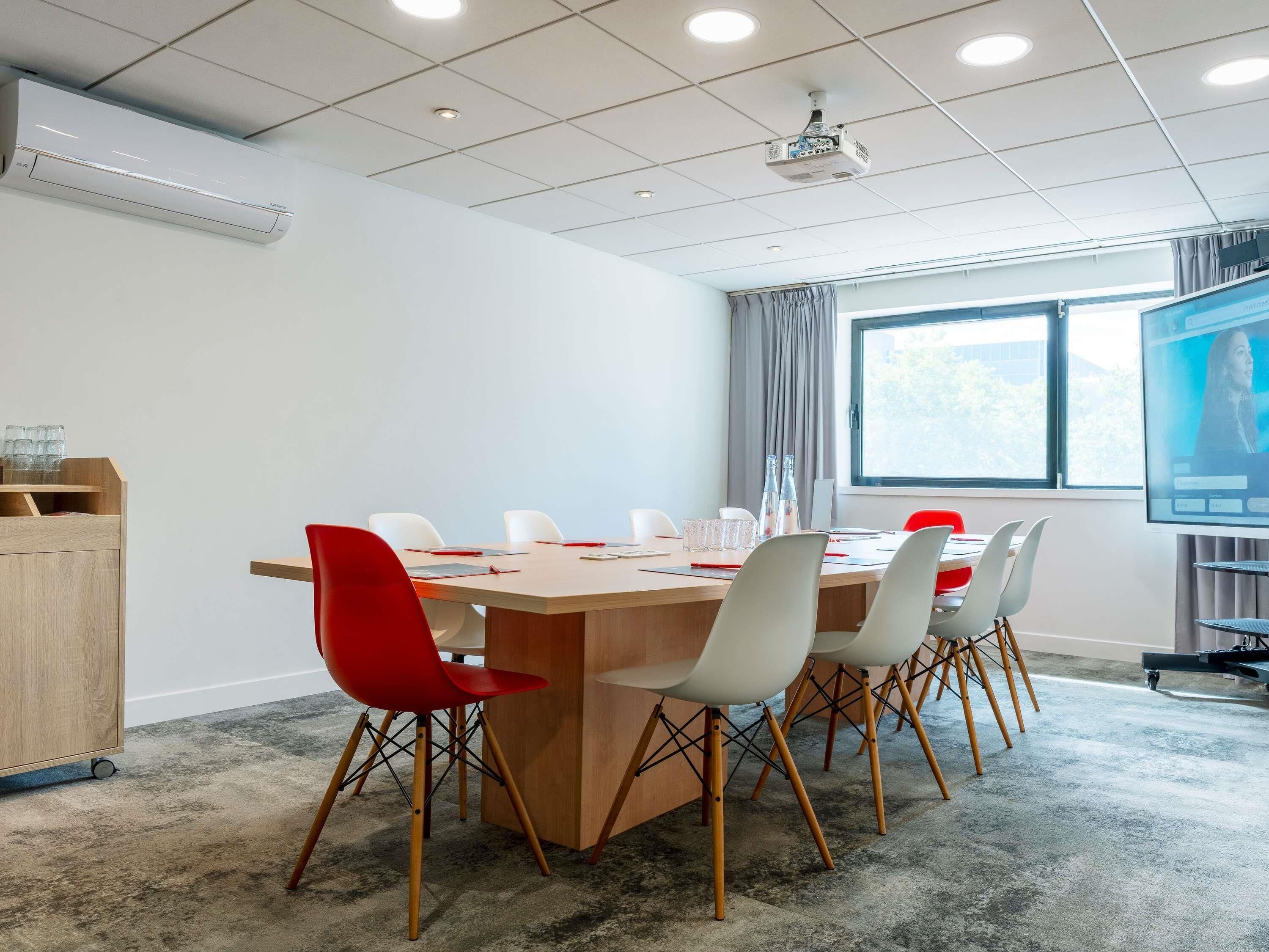 A conference room with a long table, white and red chairs, a projector screen, and a window with curtains.