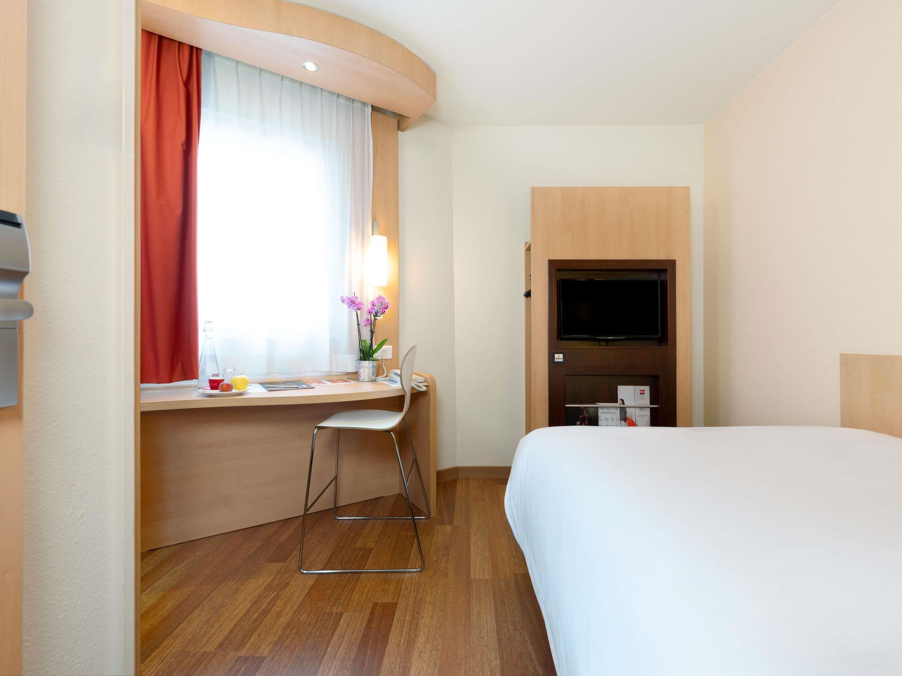 A hotel room with a bed, a desk with a chair, a TV, and a window with curtains.