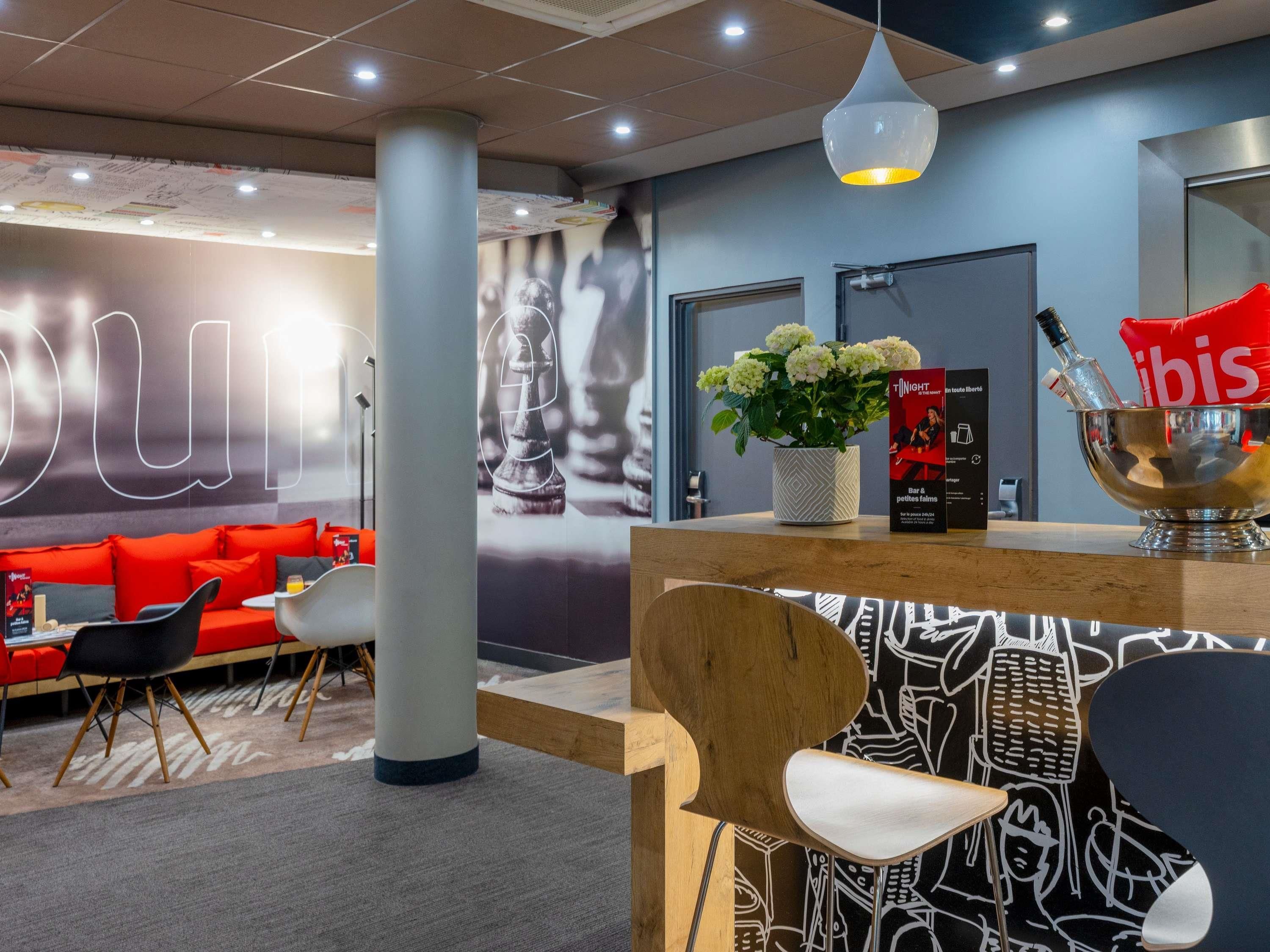 Rebedor modern d'hotel amb un sofà vermell, una taulell de recepció de fusta, una planta en test i una paret amb un disseny de peces d'escacs.