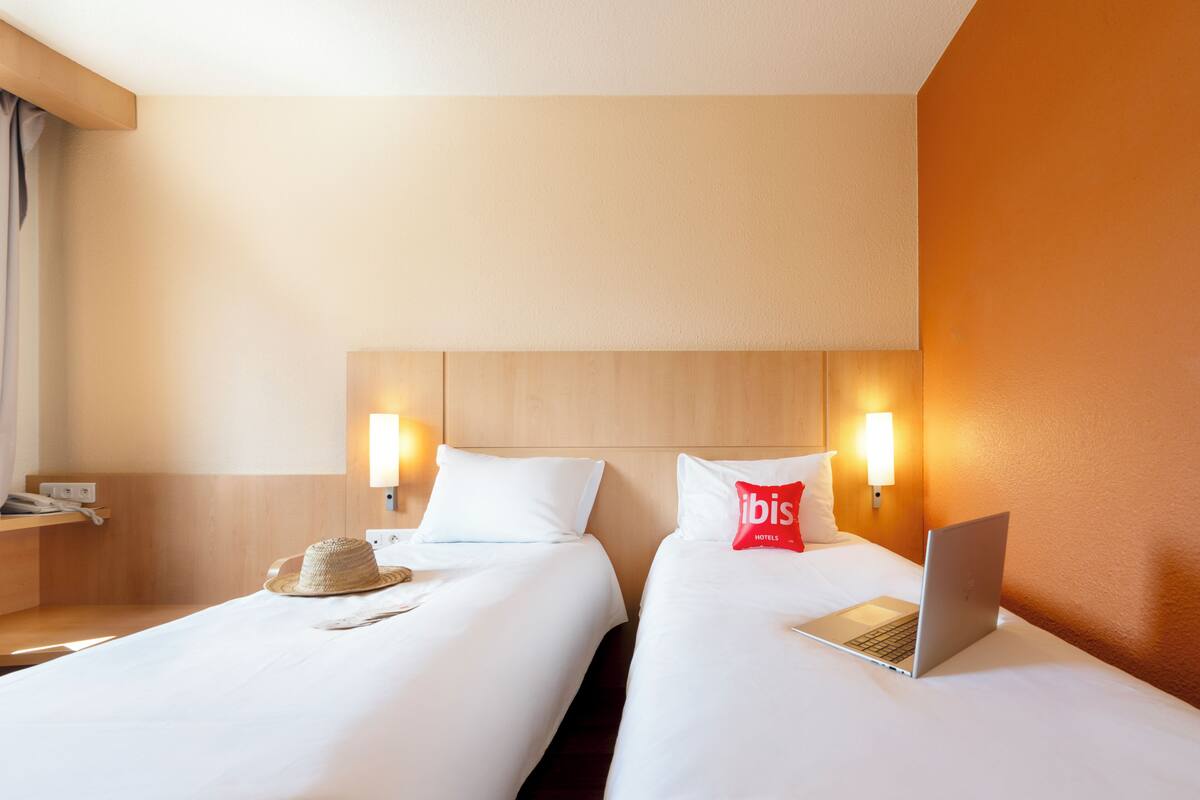 A hotel room with two beds, a laptop on one bed, a hat on the other, and a red pillow with 'ibis Hotels' branding.