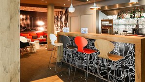 A bar area with a wooden counter, black and white patterned wall, and a seating area with red and white chairs. - ibis Nancy Centre Gare et Congres (Nancy)