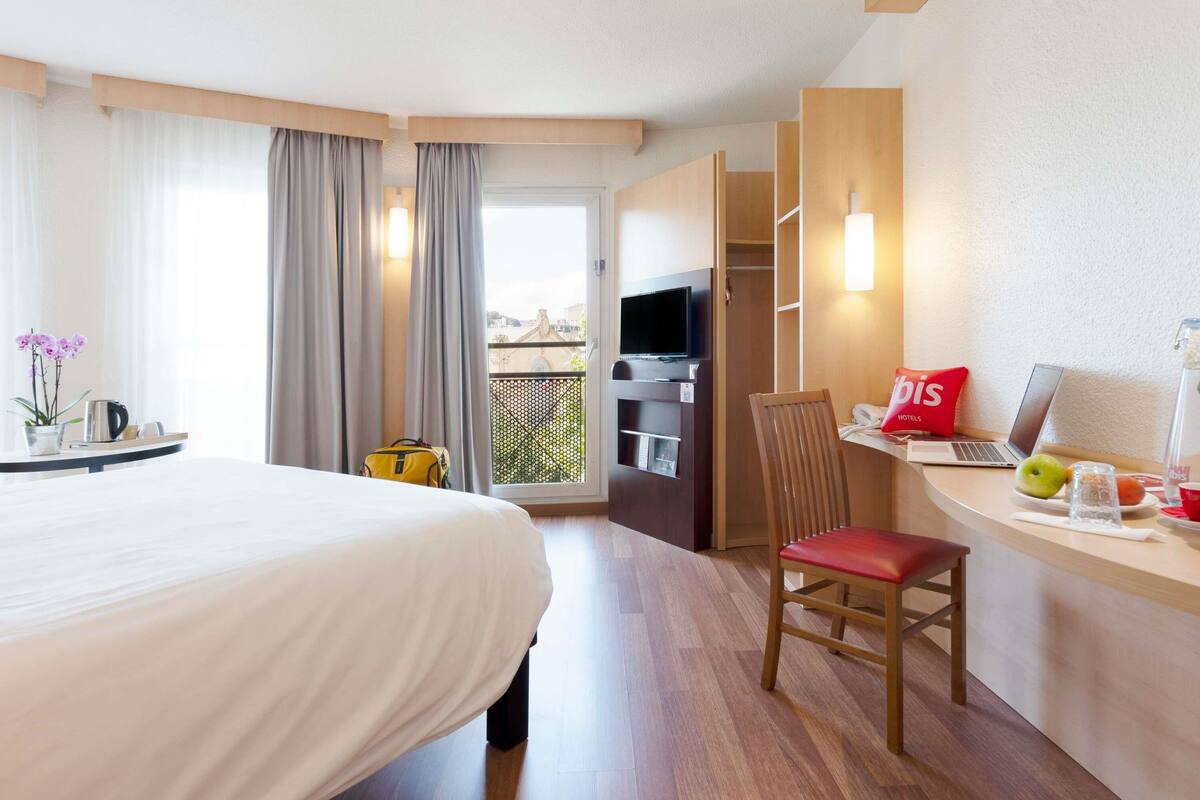 A hotel room with a bed, a desk with a laptop, a chair, a TV, and a balcony with curtains.