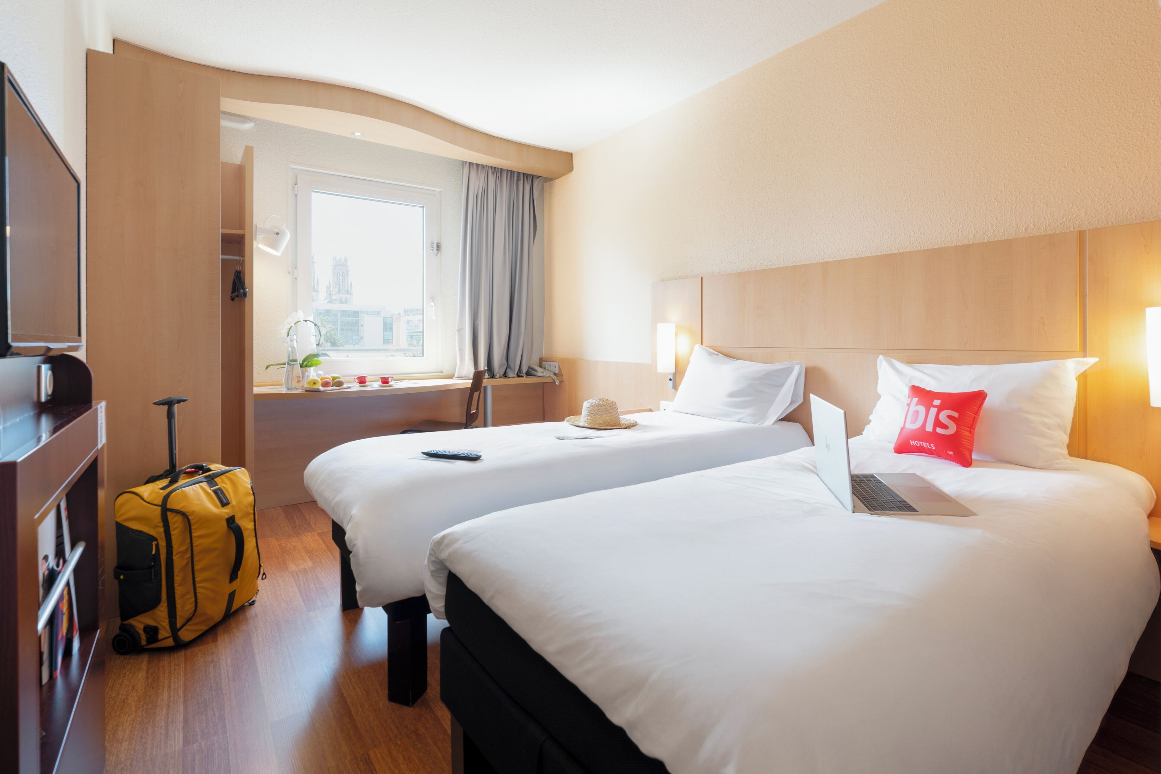 Een hotelkamer met twee bedden, een koffer, een laptop en een raam met uitzicht op gebouwen.