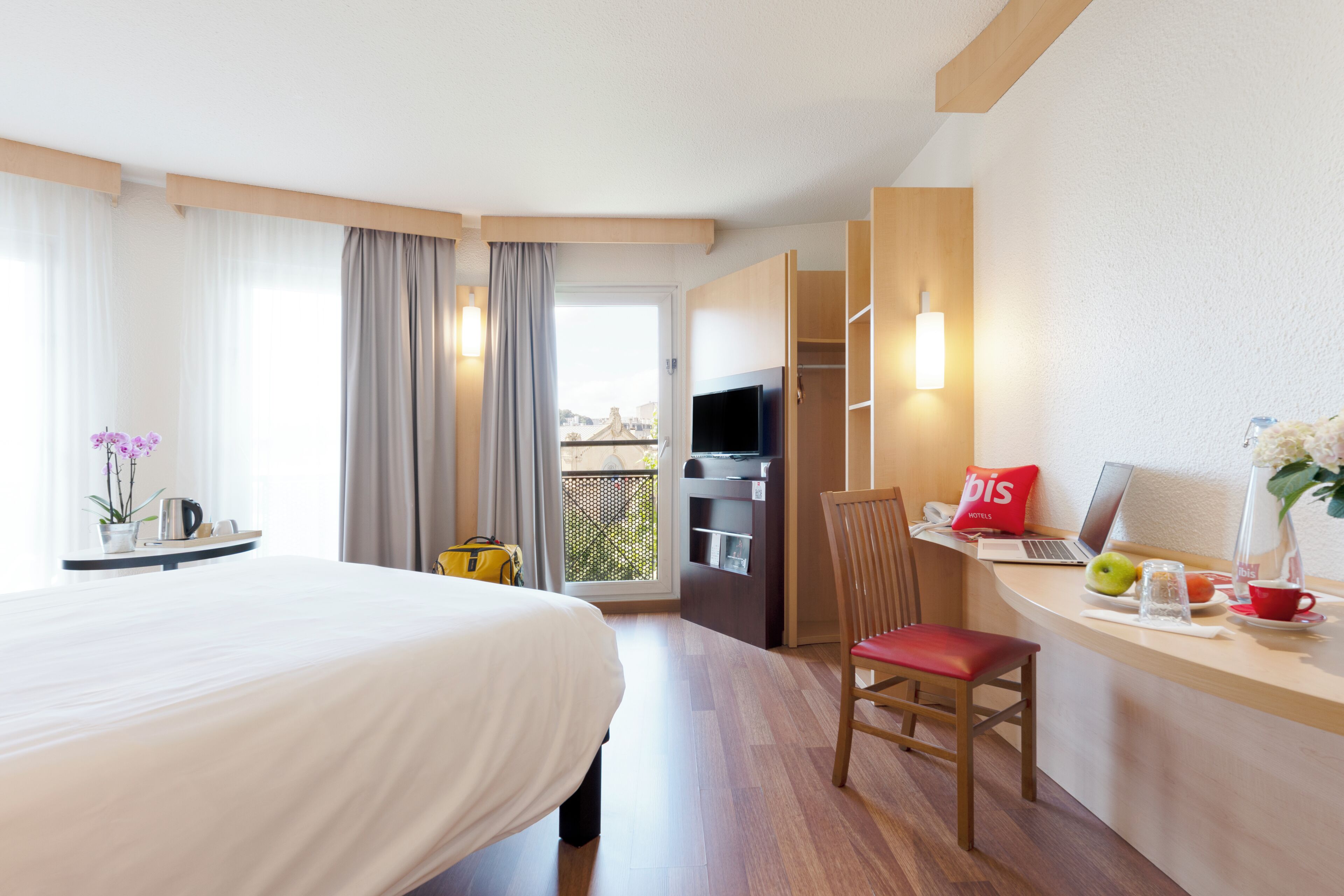 A hotel room with a bed, a desk with a laptop, a chair, a TV, and a balcony with curtains.