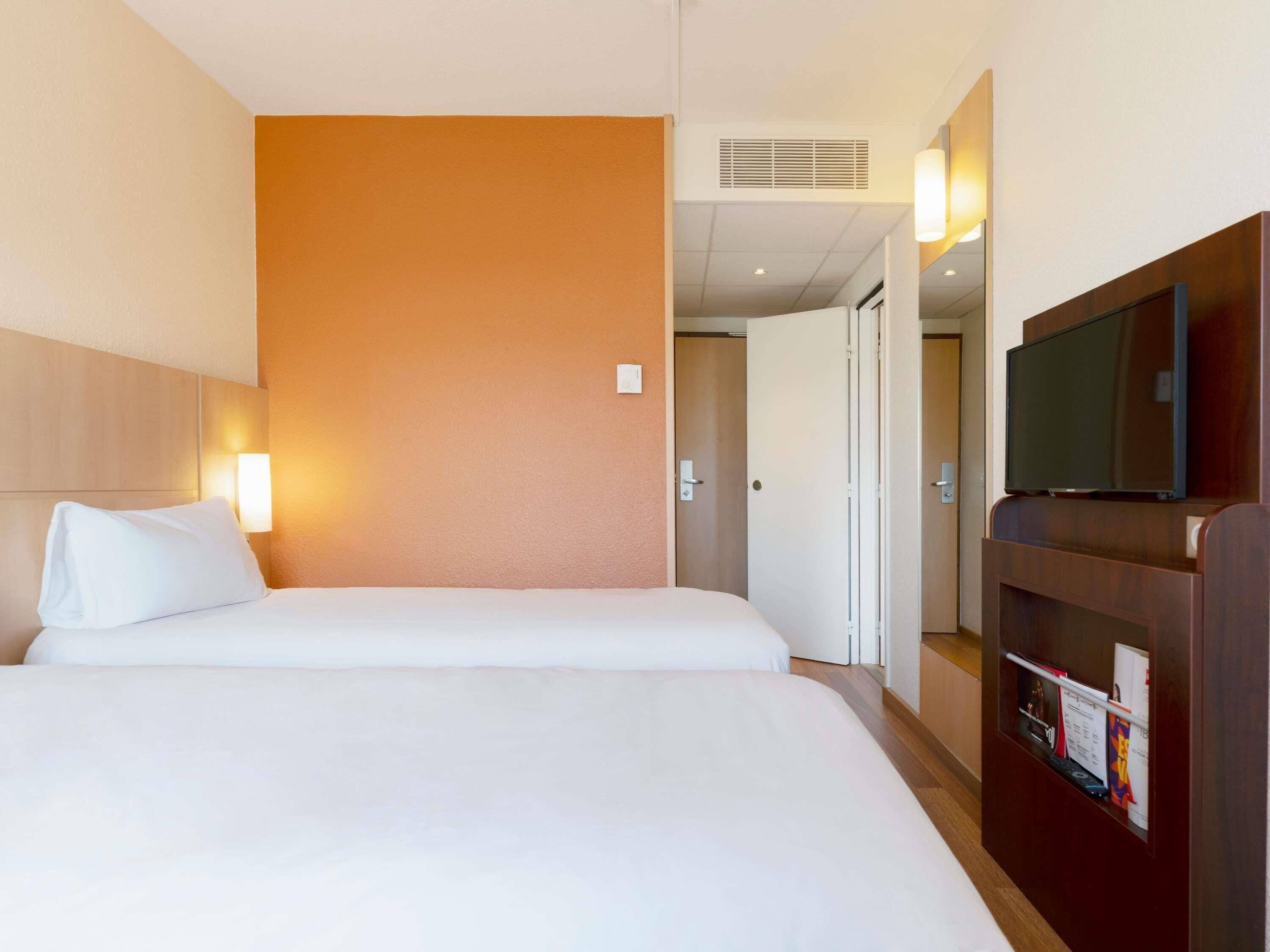 A hotel room with two beds, a television, and a wall-mounted light.