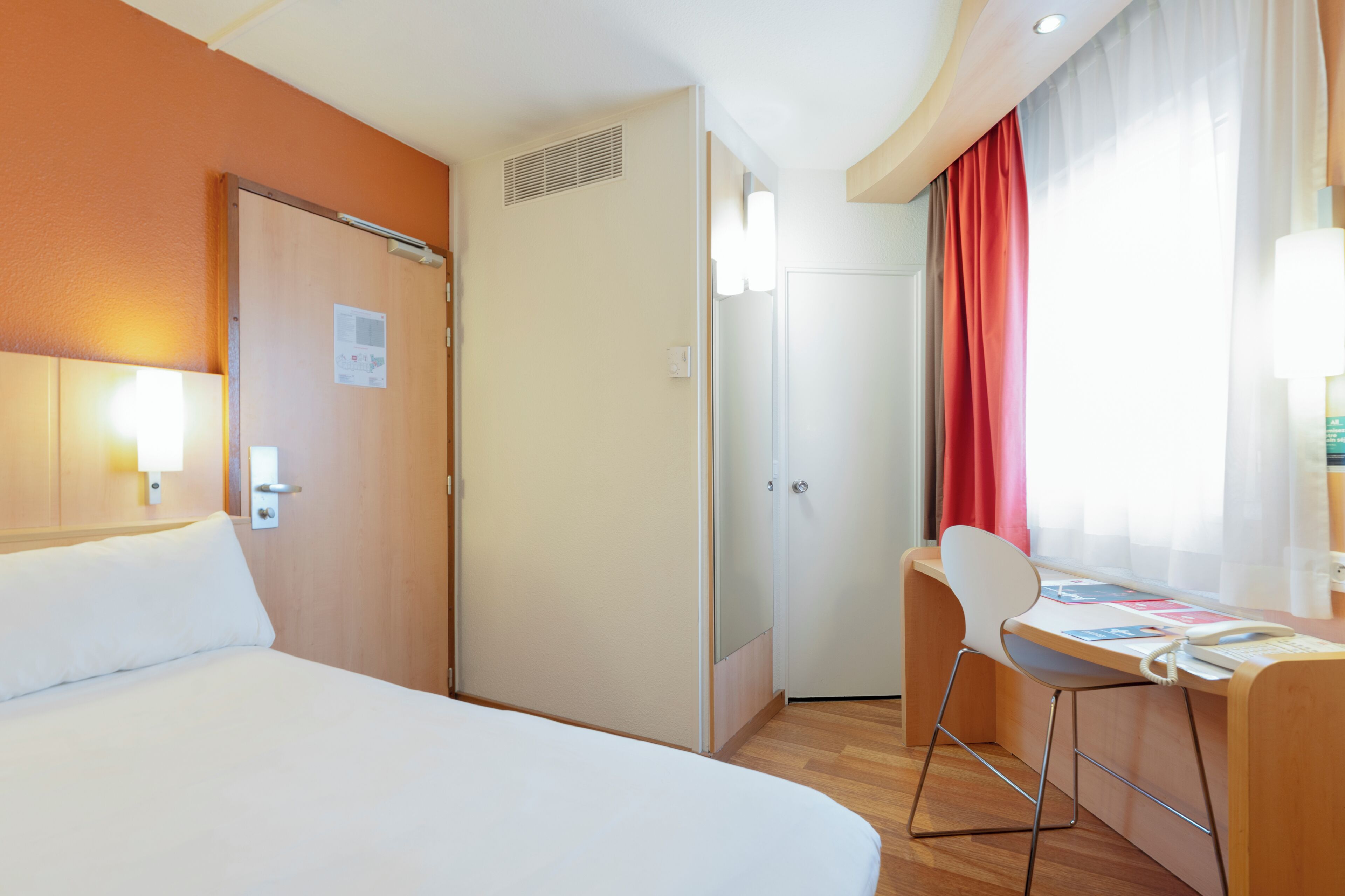 Een hotelkamer met een bed, een bureau met stoel, een deur en een raam met gordijnen.