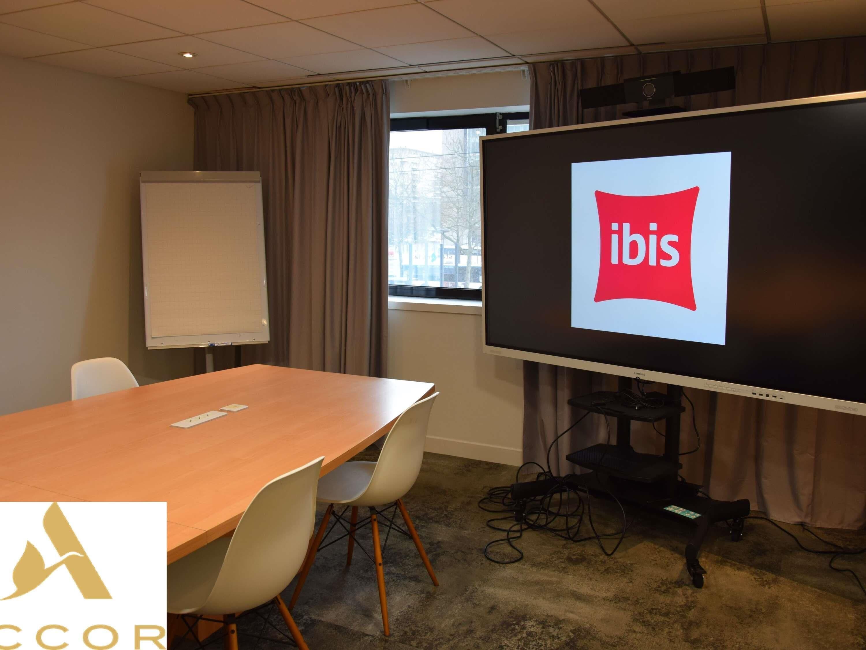 Un salón de conferencias con una pantalla grande mostrando el logotipo de ibis, una pizarra, una mesa de madera y sillas blancas.