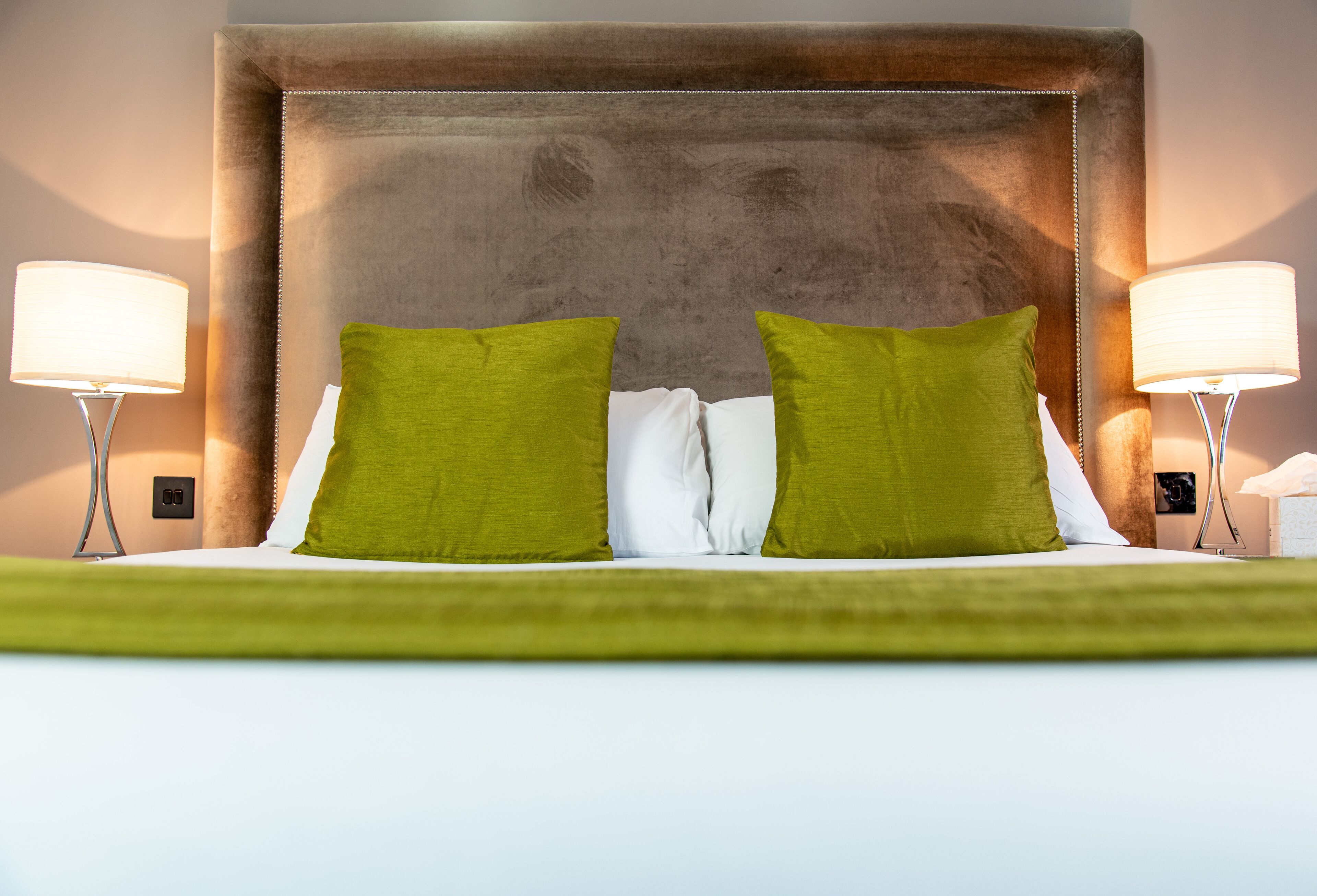 A hotel room with a large bed, green and white pillows, and two bedside lamps.