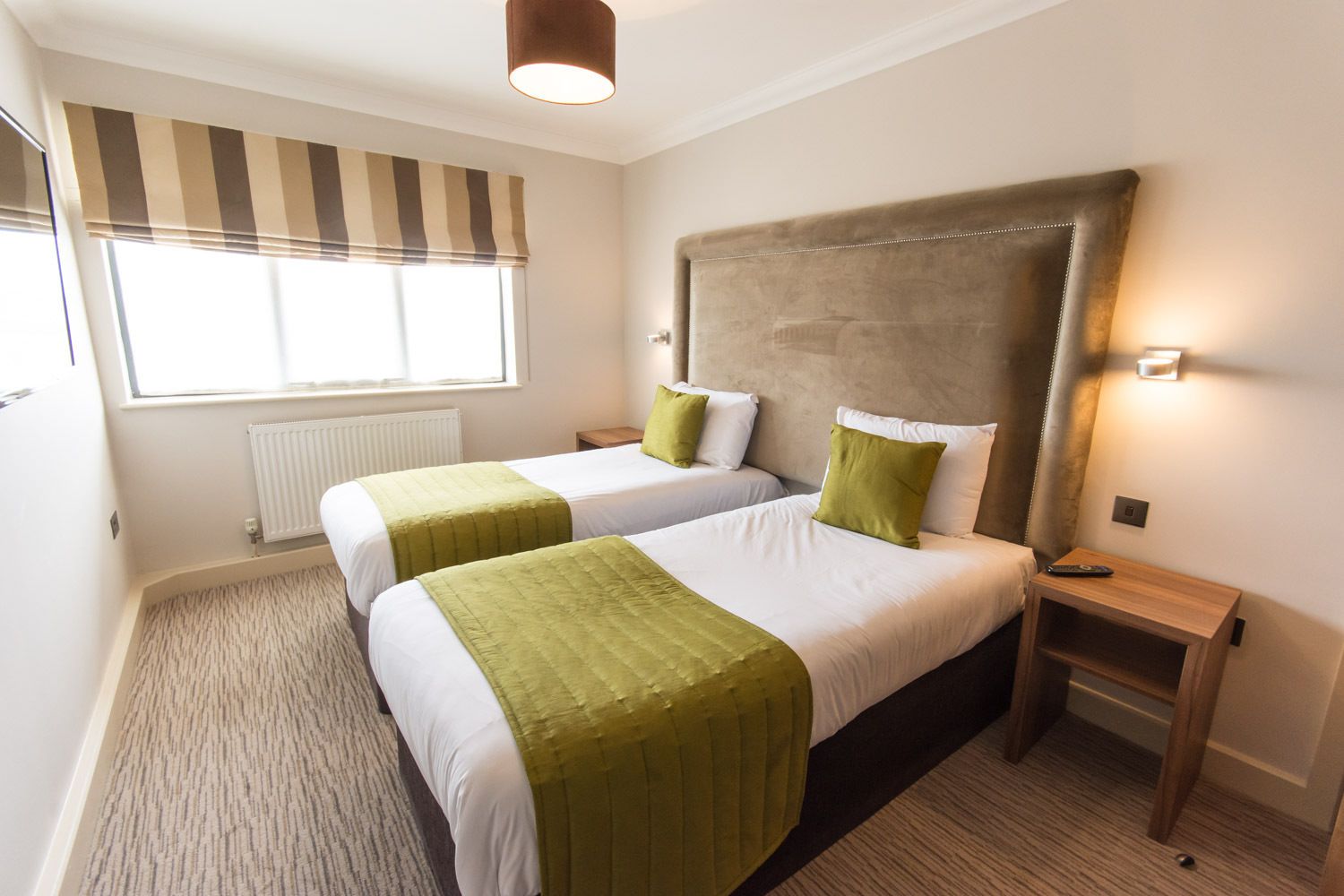 A hotel room with two beds, a large headboard, a window with striped blinds, a radiator, and a nightstand with a remote control.