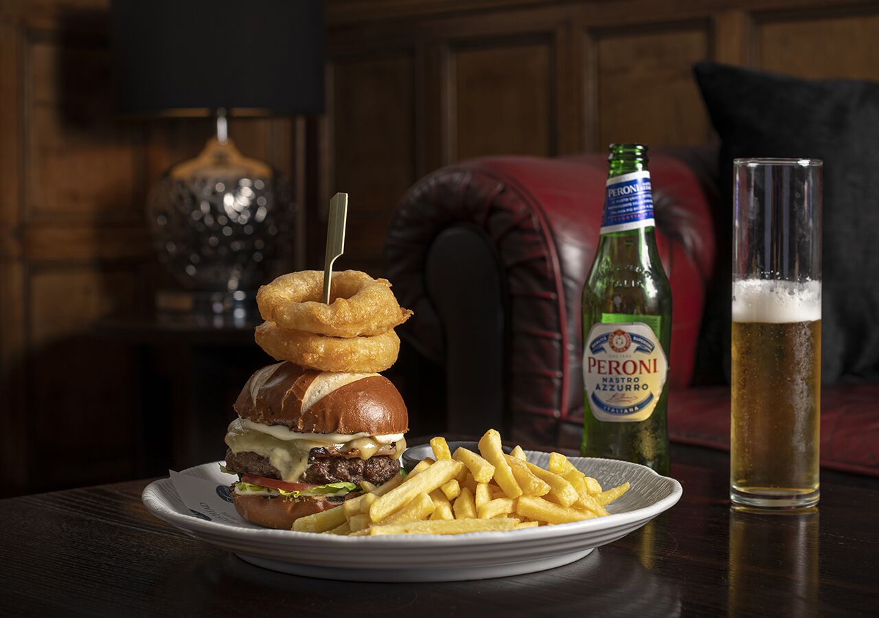 Une assiette avec un hamburger, des oignons frits et des frites, une bouteille de bière Peroni et un verre de bière sur une table.