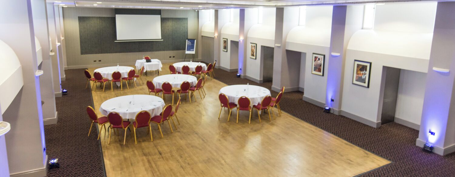 A conference room with multiple round tables and red chairs, a projector screen, and framed pictures on the walls.