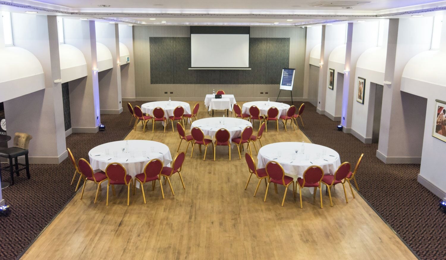 A conference room with multiple round tables and red chairs, a projector screen, and a whiteboard.