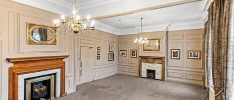 A spacious room with a fireplace, chandeliers, and framed pictures on the walls.