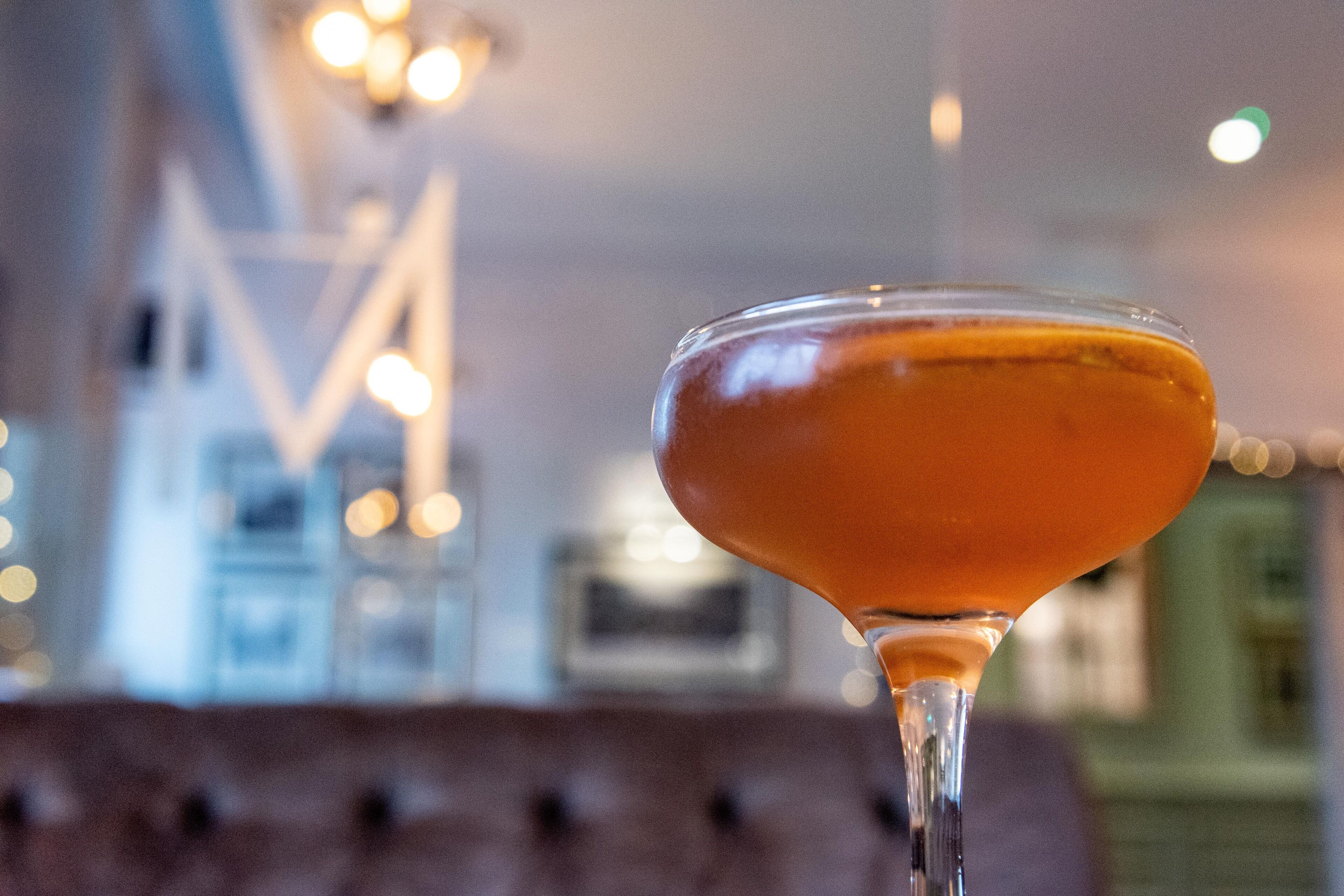 Un verre à martini contenant un liquide transparent, vraisemblablement une boisson alcoolisée, dans un intérieur bien éclairé, avec un arrière-plan flou où l’on aperçoit un grand luminaire abstrait.