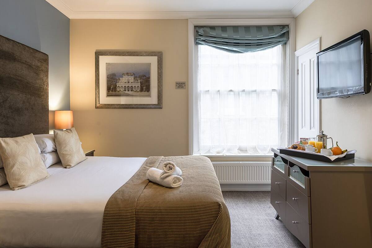 Ein Hotelzimmer mit Bett, Schreibtisch und Frühstücksgebäck sowie einem Bild an der Wand und einem mit Vorhängen versehenen Fenster.