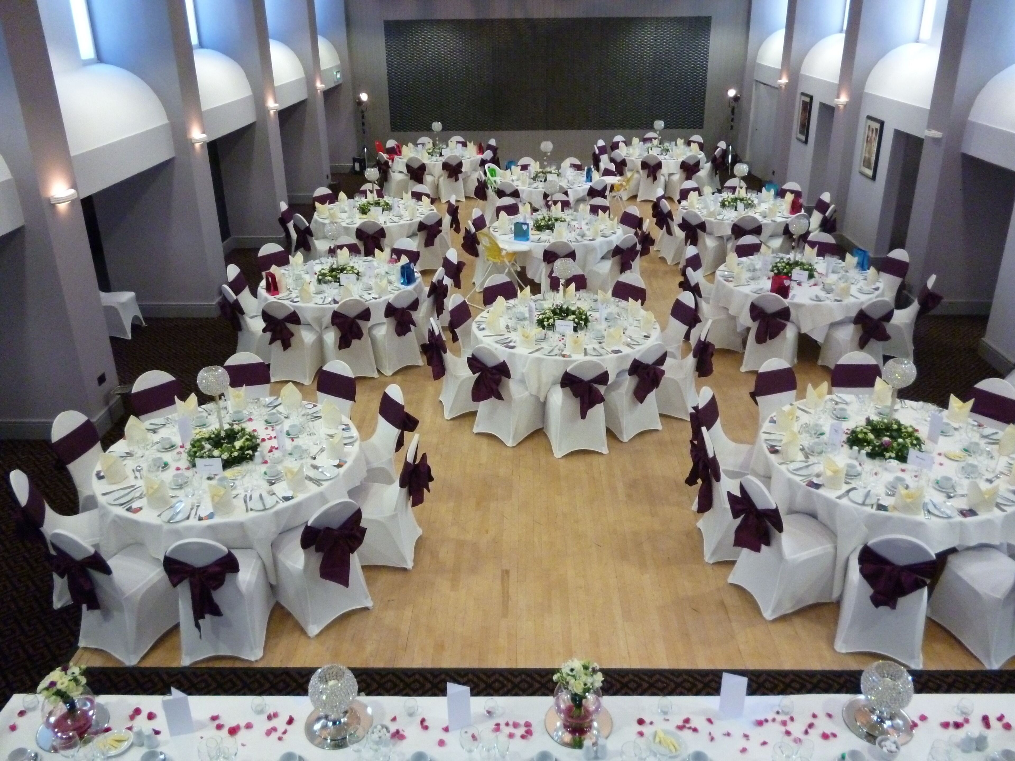 Une salle de banquet avec de nombreuses tables rondes, chacune recouverte d’une nappe blanche et ornée d’un ruban violet.