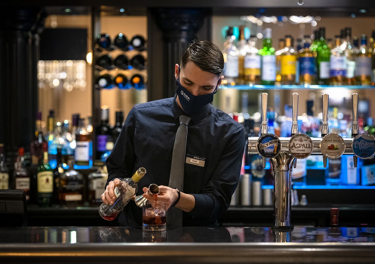 A bartender preparing a drink at a bar with multiple liquor taps and a well-stocked shelf of bottles in the background.