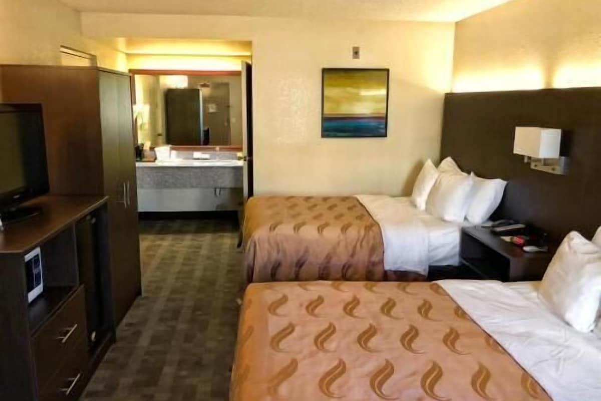 A hotel room with two beds, a TV, a desk, and a bathroom visible through an open door.