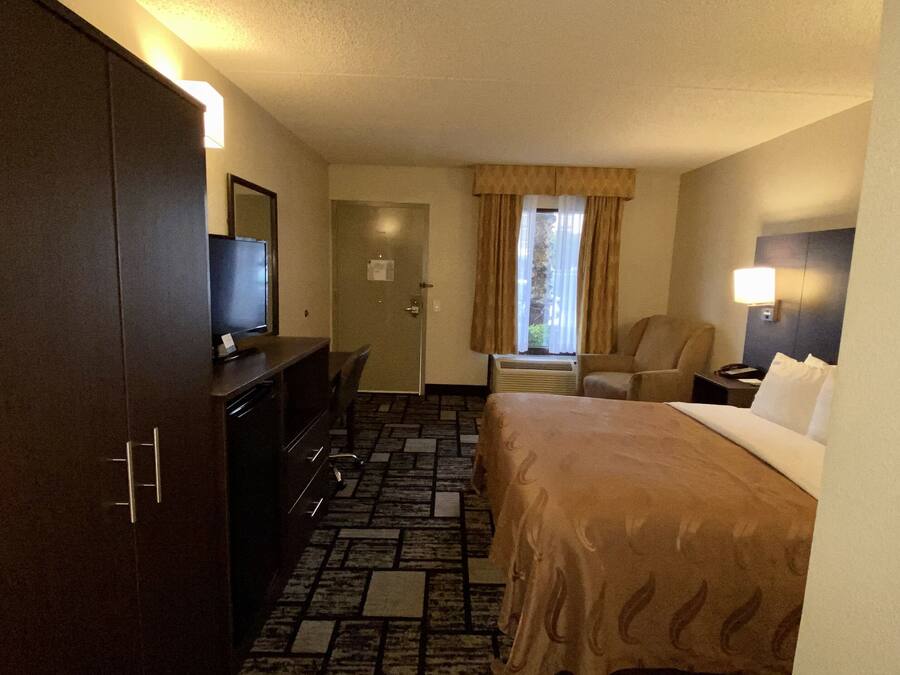 Habitación de hotel con cama, escritorio con televisor, silla, mesita de noche y ventana con cortinas.