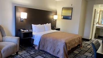 Habitación de hotel con cama, una silla, un escritorio, una mesita de noche, una lámpara y un cuadro en la pared.