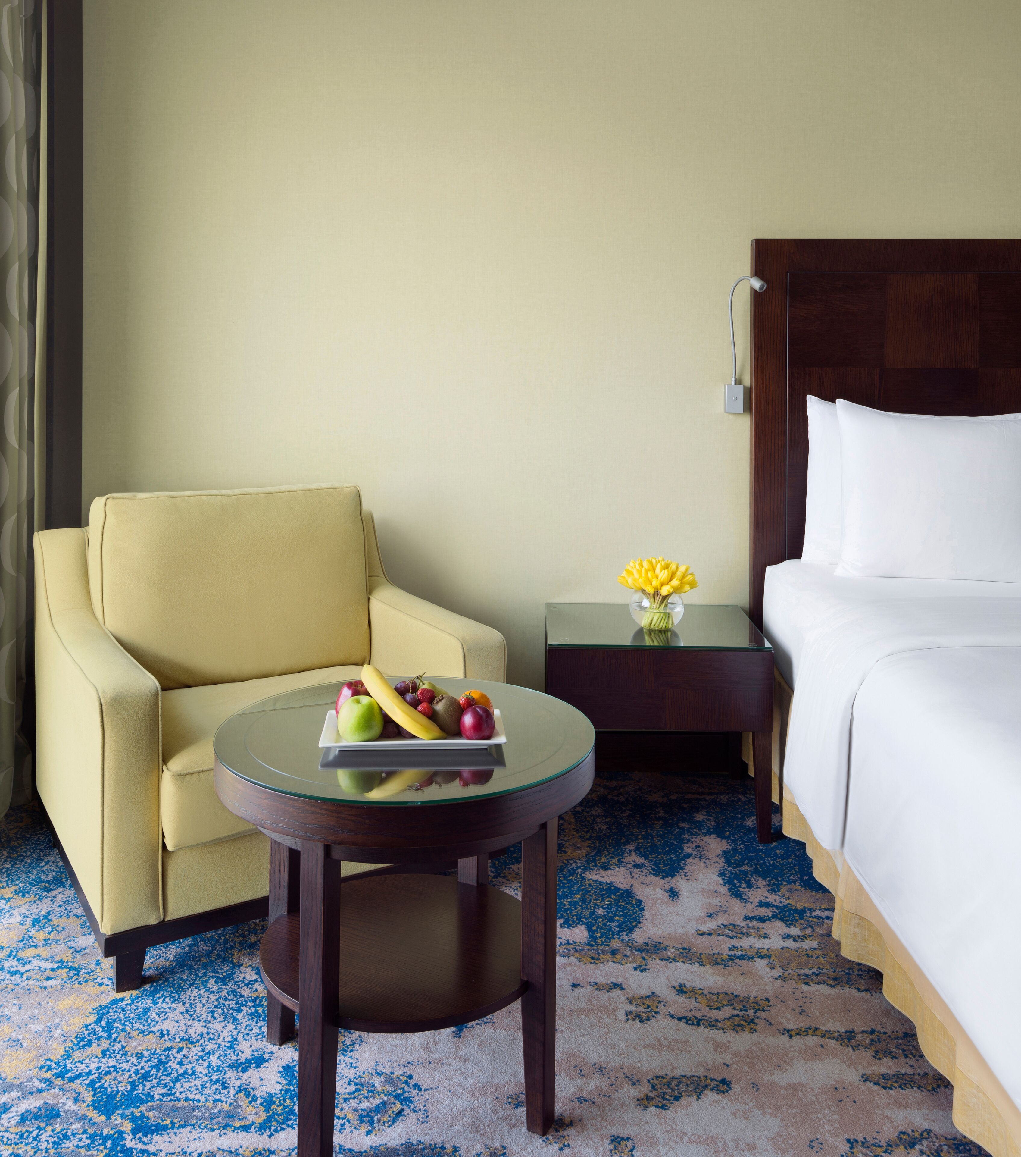 Habitación de hotel con cama, una silla, una mesita con fruta y una mesita de noche con un florero.