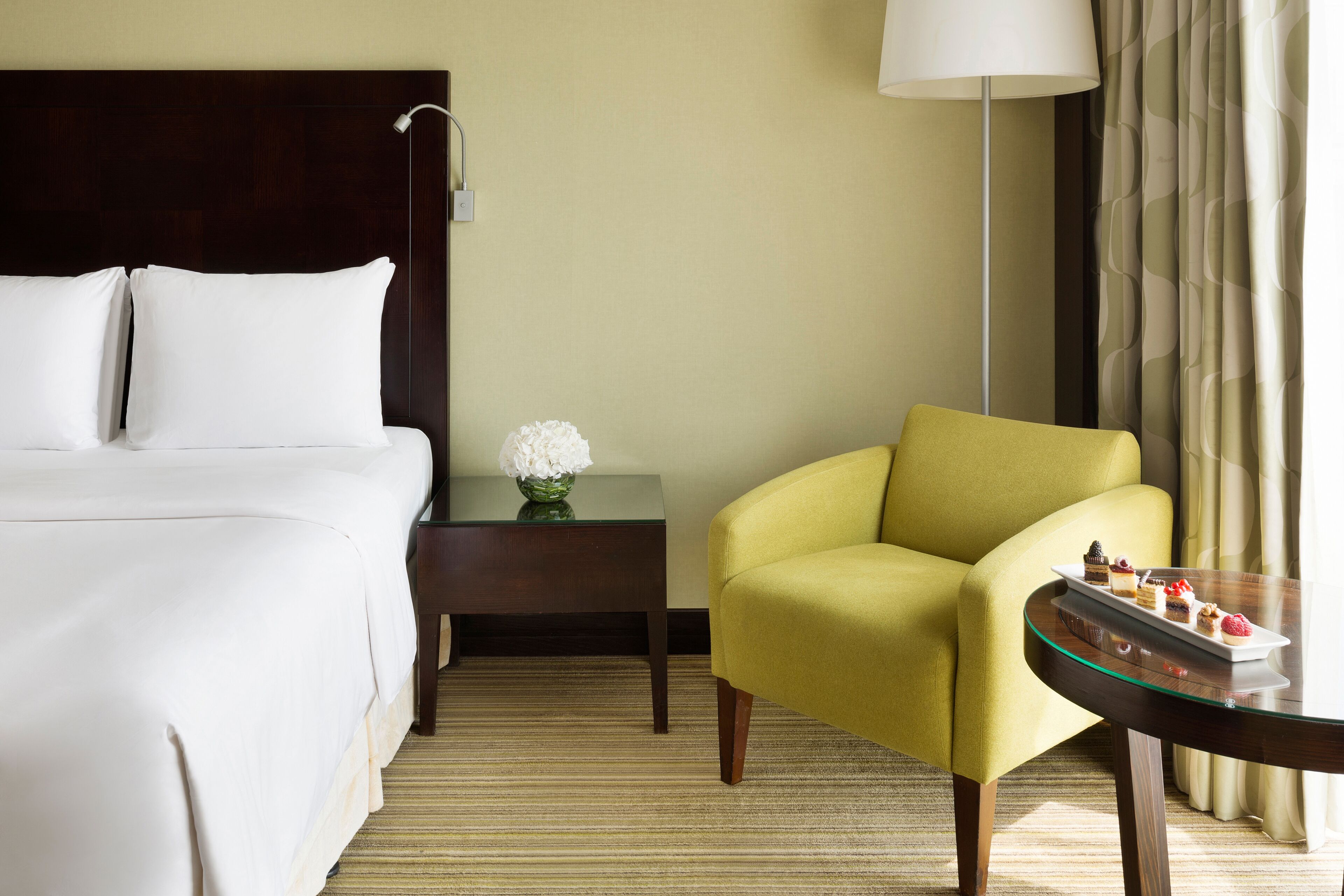 A hotel room with a bed, a nightstand with a vase, a yellow armchair, and a table with desserts.