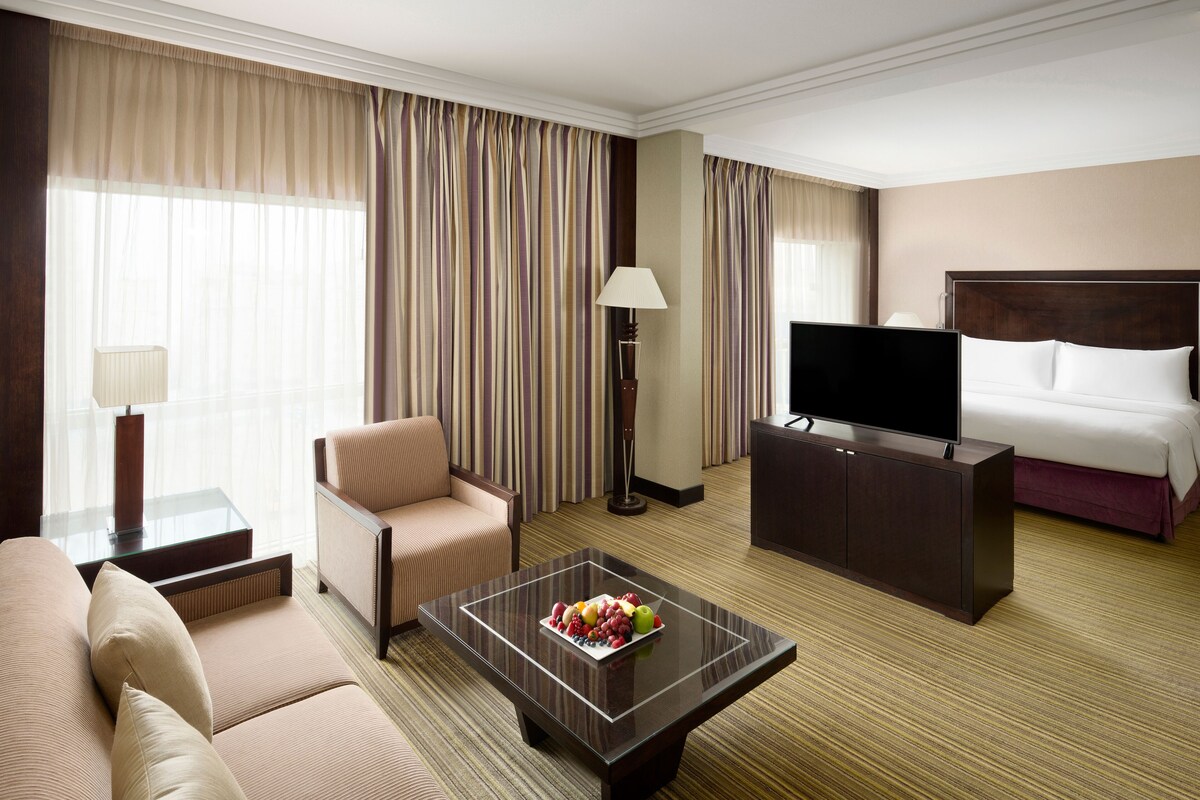 Une chambre d’hôtel équipée d’un canapé, d’un fauteuil, d’une télévision, d’une table de chevet et d’un panier de fruits posé sur une table basse.
